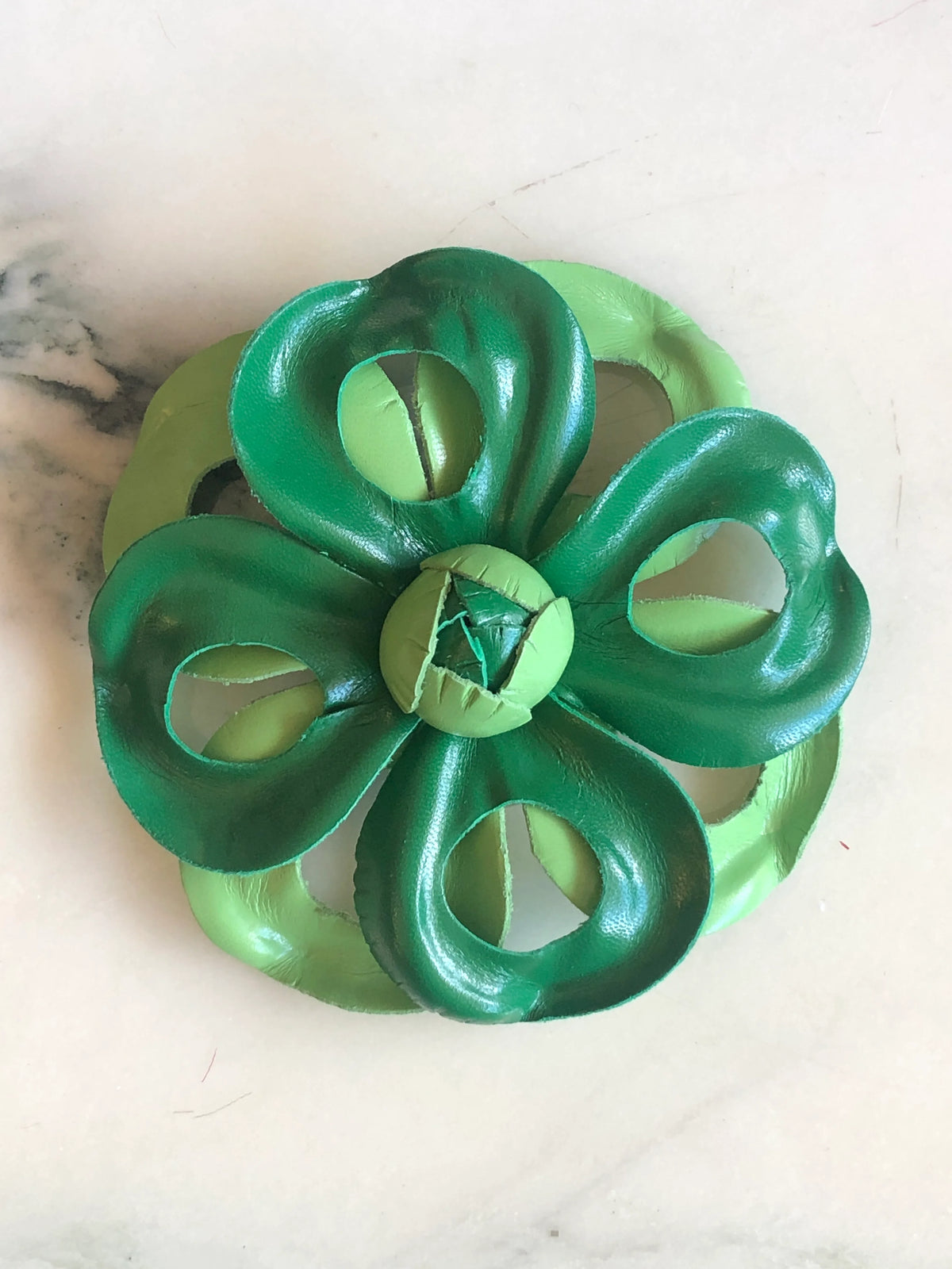 Broochella Viola del Pensiero Leather Flower Brooch Pin in green, featuring sculptural two-tone dark and light green leather petals with open cut-out detailing and a handcrafted centre on a cream marble background.
