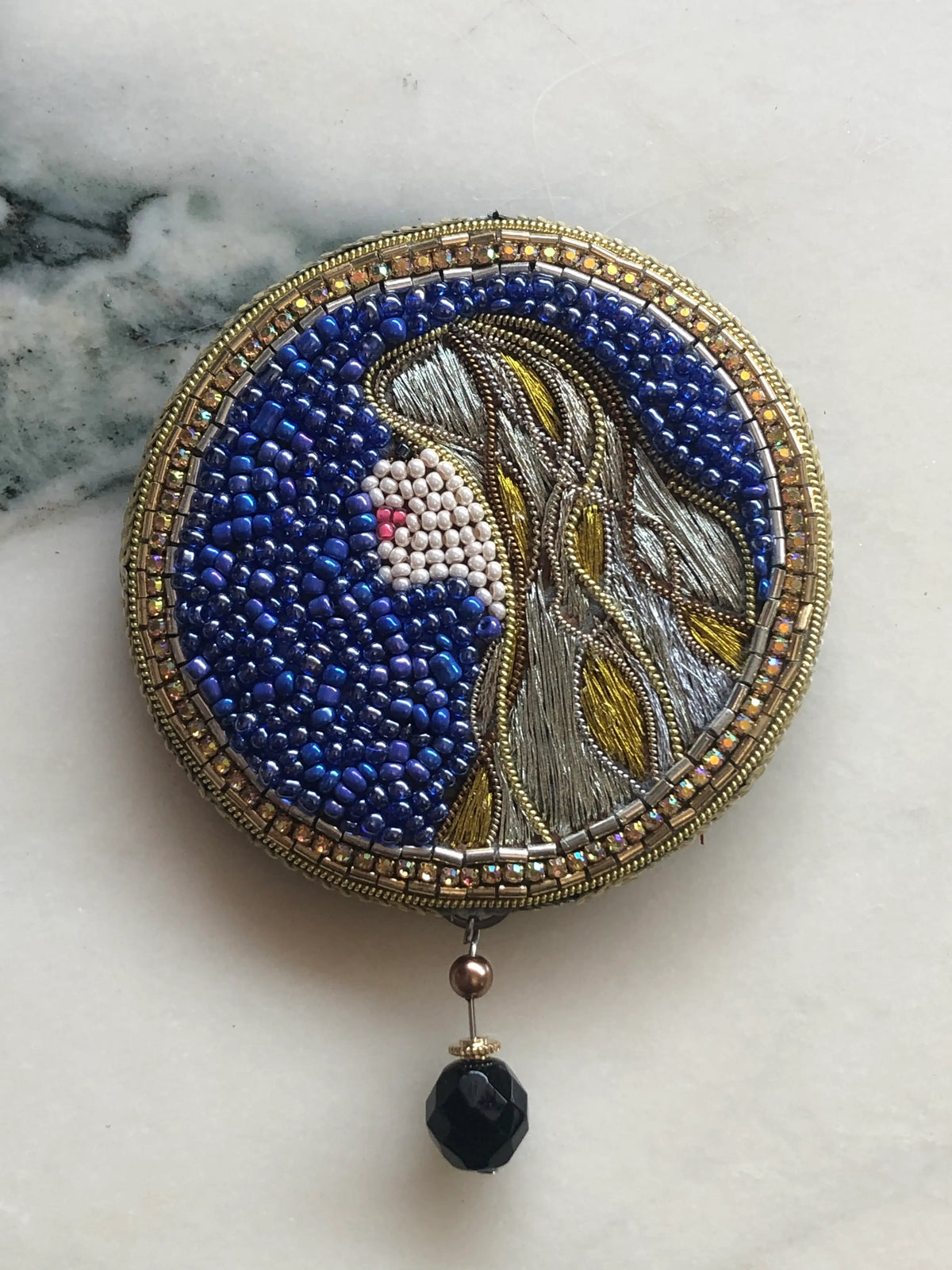 A Broochella hand beaded Virgo Zodiac  round beaded brooch featuring a stylized profile portrait of a woman with blue, silver, and gold beadwork, resting on a white marble surface with a black bead dangling below.
