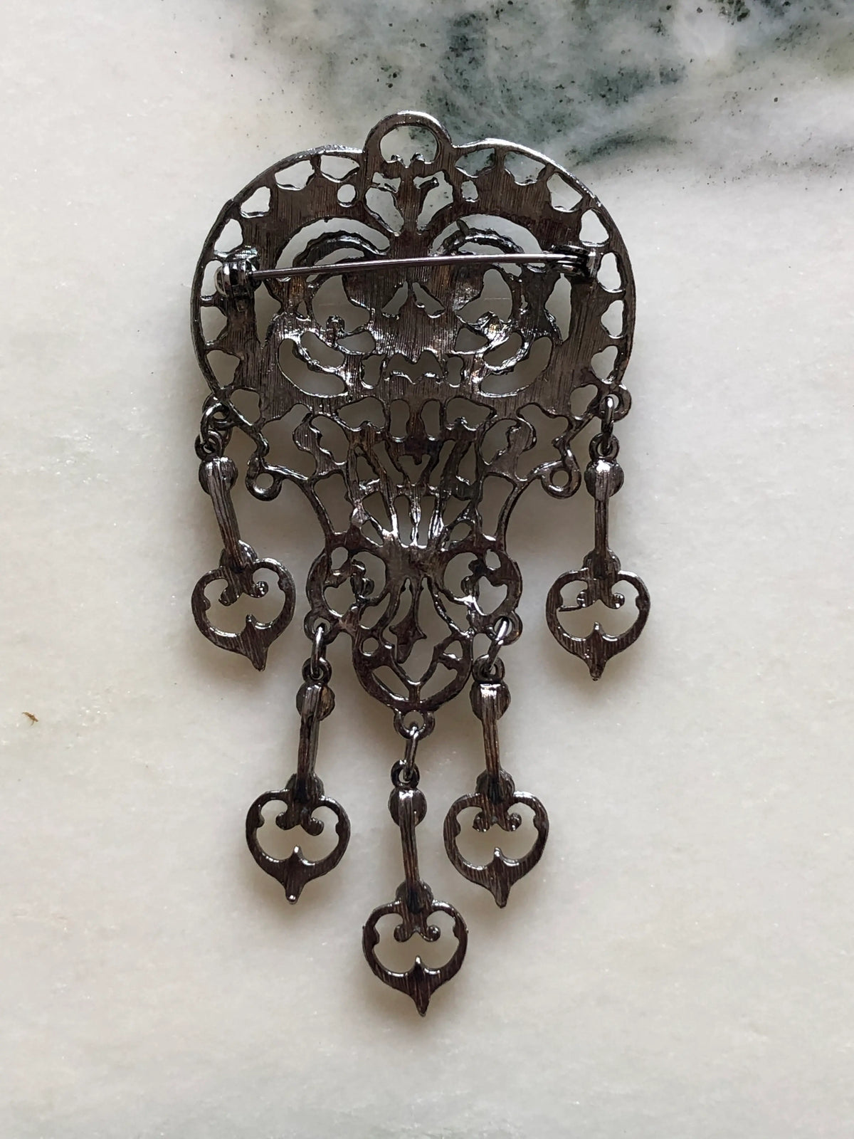 Broochella Vittoria Vintage Silver Filigree Drop Brooch featuring ornate silver-tone openwork, five articulated drops and green, red and clear stones on a cream and grey marble background.