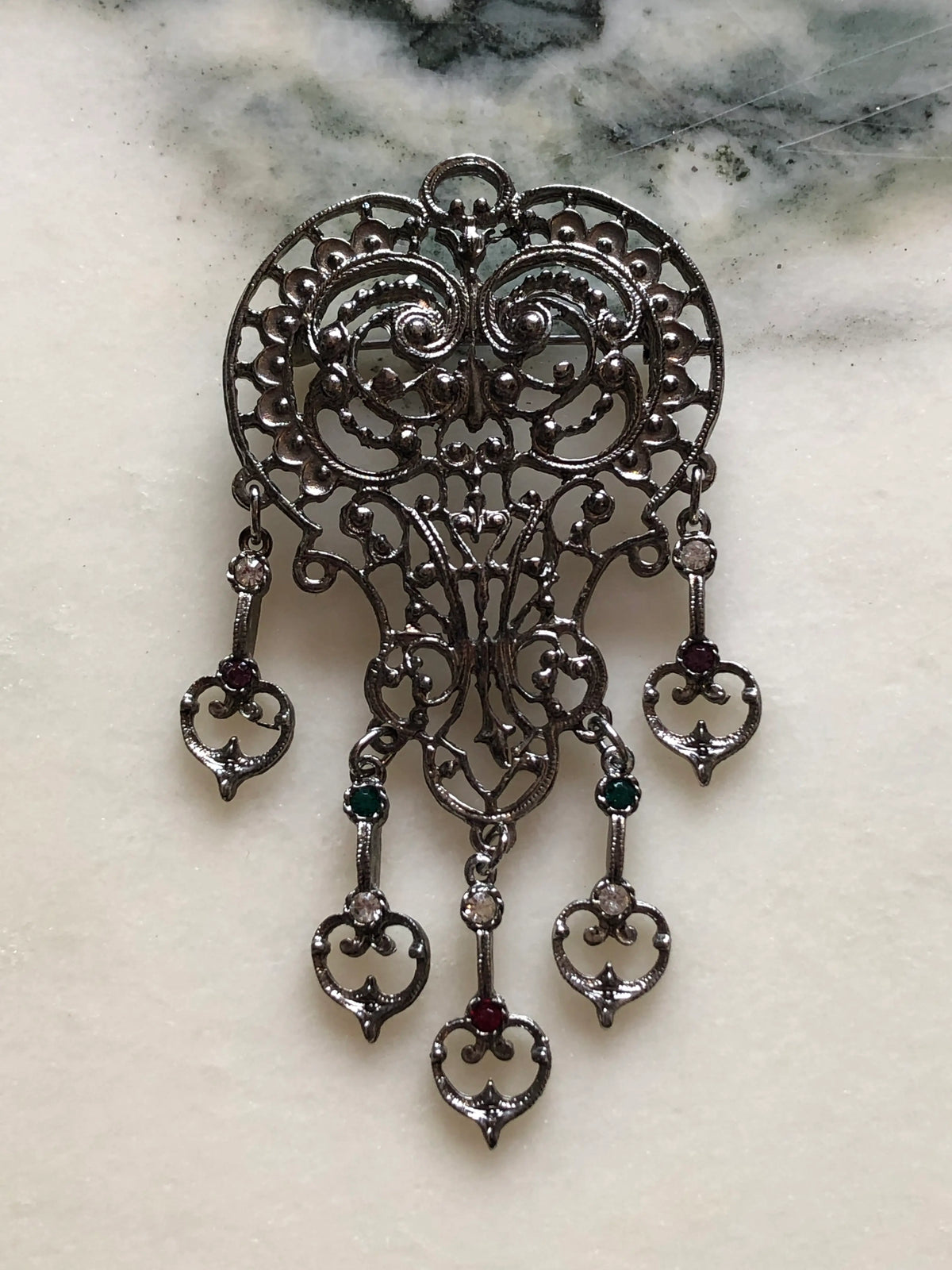 Broochella Vittoria Vintage Silver Filigree Drop Brooch featuring ornate silver-tone openwork, five articulated drops and green, red and clear stones on a cream and grey marble background.
