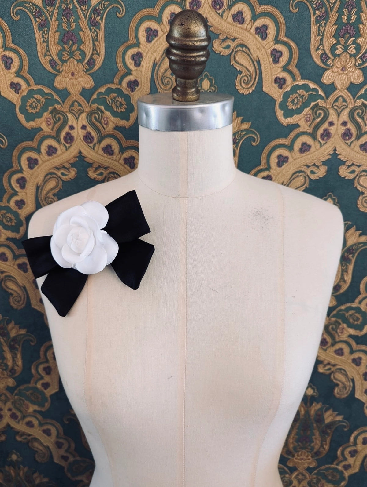 Broochella viva l'amor handmade silk black bow brooch with white flower centre.  It is pinned to a mannequin. The background is green and gold damask wallpaper.
