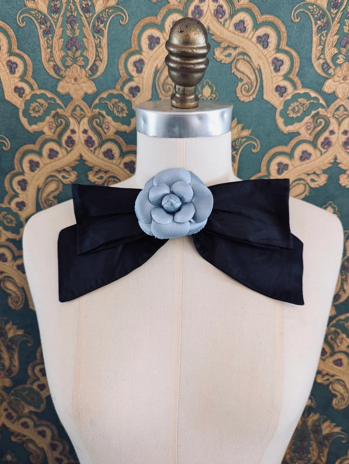 Broochella viva l'amor handmade black silk bow brooch, with blue flower embellishment.  The background is green and gold damask wallpaper.