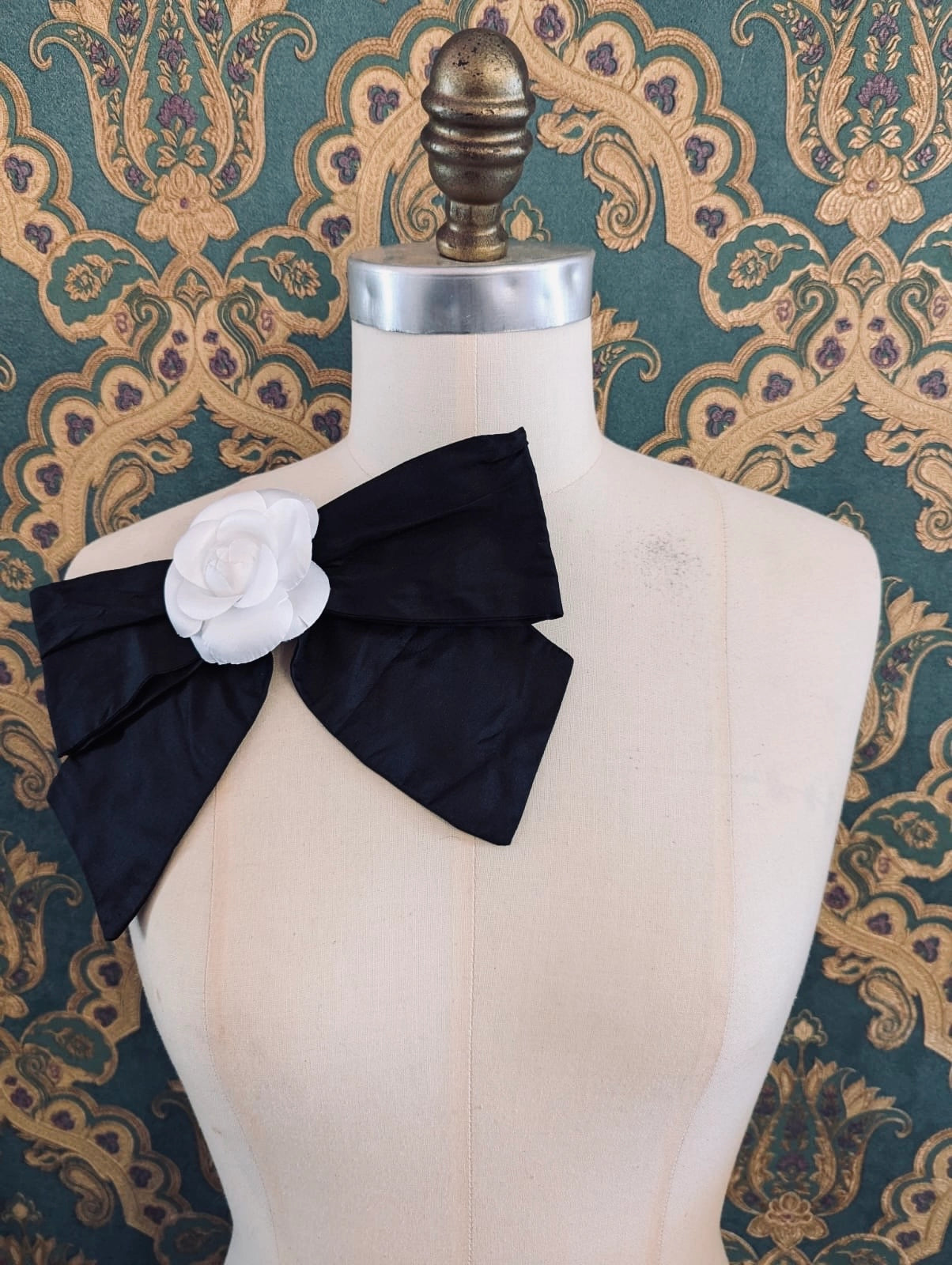 Broochella viva l'amor handmade black silk bow brooch, with white flower embellishment.  The background is green and gold damask wallpaper.