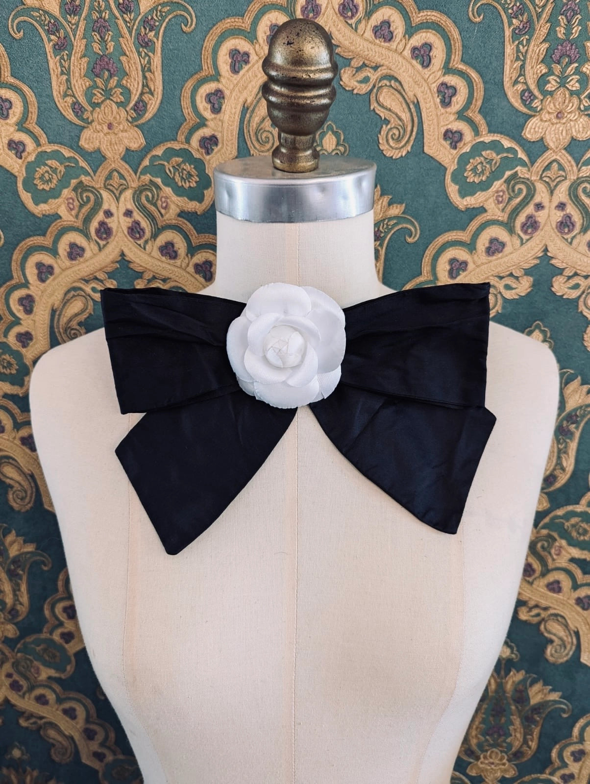 Broochella viva l'amor handmade black silk bow brooch, with white flower embellishment.  The background is green and gold damask wallpaper.