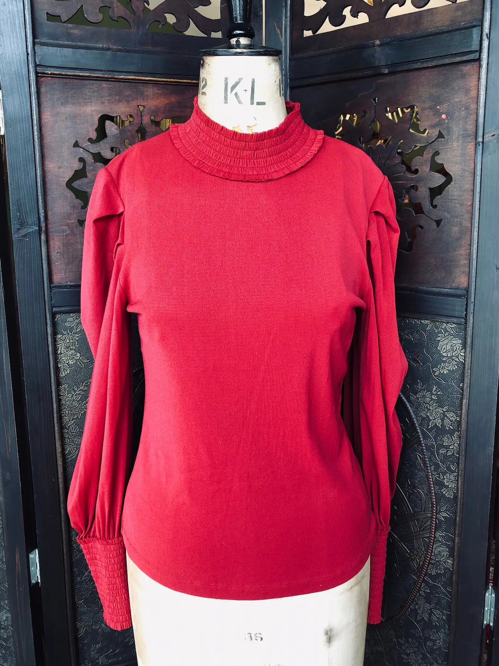 Broochella The Stylist's Closet Vivienne Tam red, long sleeve mesh top. It has pleated shoulder details and shirring at the wrists and neck. It is pictured on a vintage mannequin. The background is an ornate, vintage, dark wood room divider. 