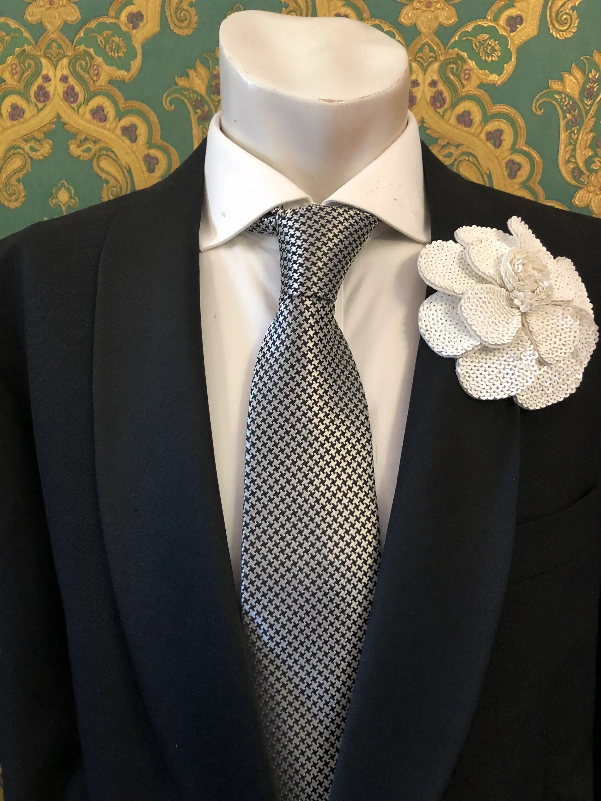 A men's formal accessory set displayed on a white mannequin torso, featuring a black suit jacket, a white dress shirt, a silver and black checkered tie, and an orange Men's Broochella Scintillante sequinned flower brooch lapel pin against a green and gold patterned wallpaper background.