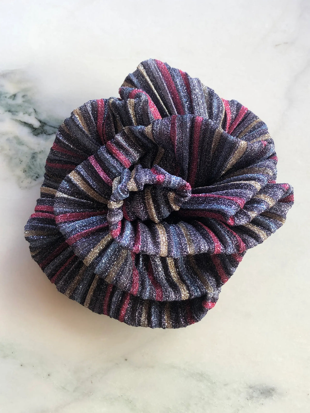 Broochella Wonka Brooch in black metallic pleated fabric with red, silver and gold stripes, sculpted into a statement flower on a white marble background.
