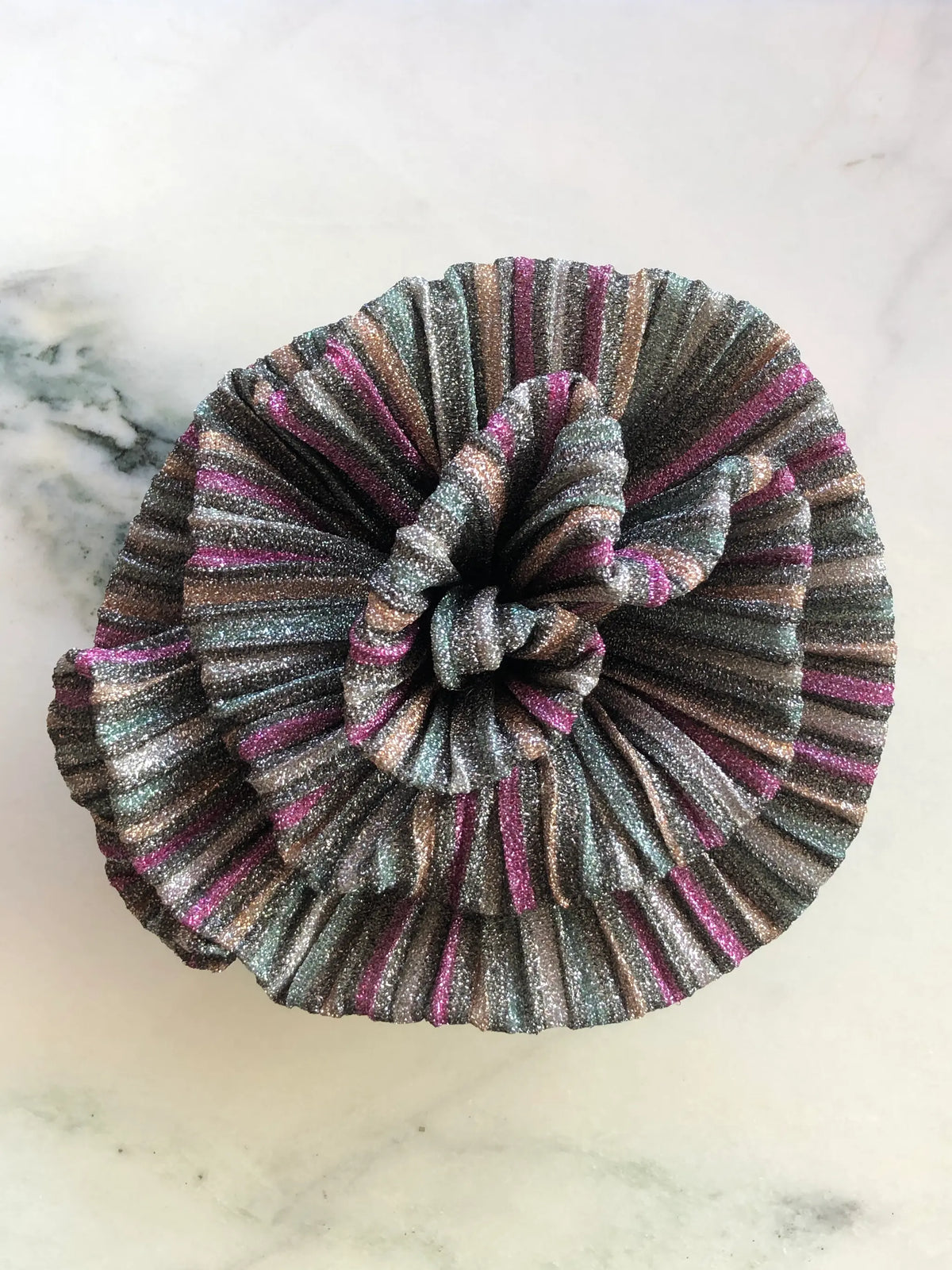 Broochella Wonka Brooch in shimmering green pleated fabric with pink, silver and bronze stripes, sculpted into a statement flower on a white marble background.