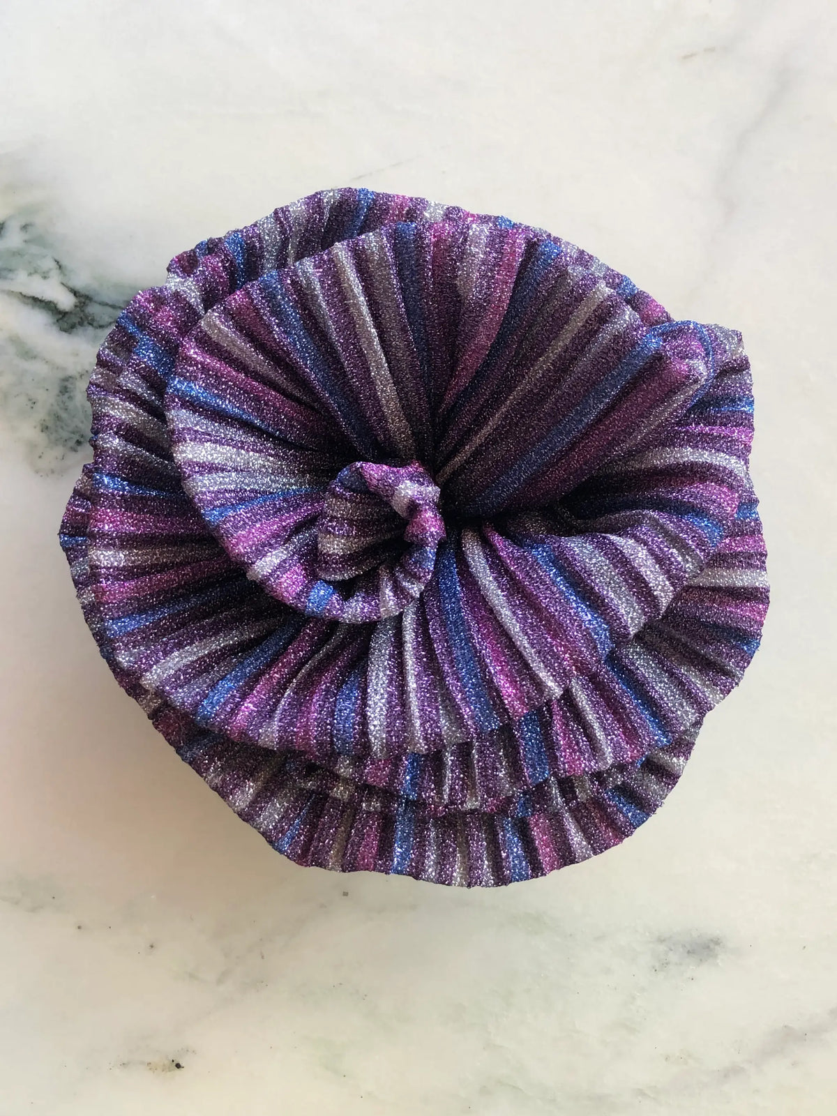 Broochella Wonka Brooch in shimmering purple pleated fabric with blue, pink and silver stripes, sculpted into a statement flower on a white marble background.