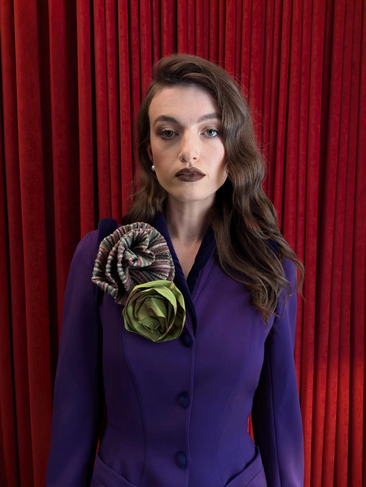 A woman with wavy brown hair and dark lipstick stands in front of a red curtain backdrop, wearing a purple tailored jacket or dress with a large Broochella wonka  fabric flower brooch embellishment on the chest in olive green and brown tones.