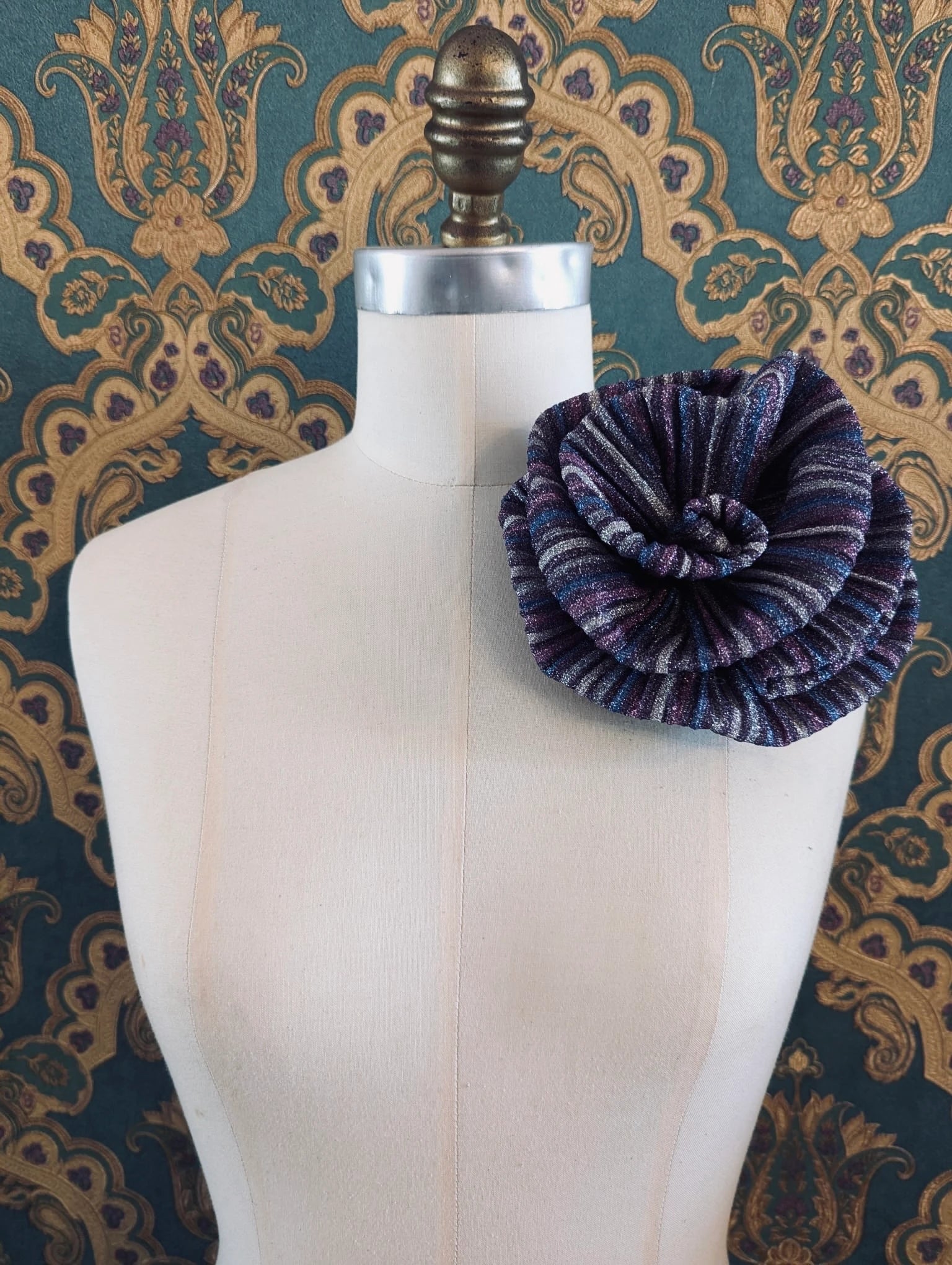 Broochella wonka handmade flower brooch in blue purple striped lurex. It is pinned to a mannequin. The background is green and gold damask wallpaper.