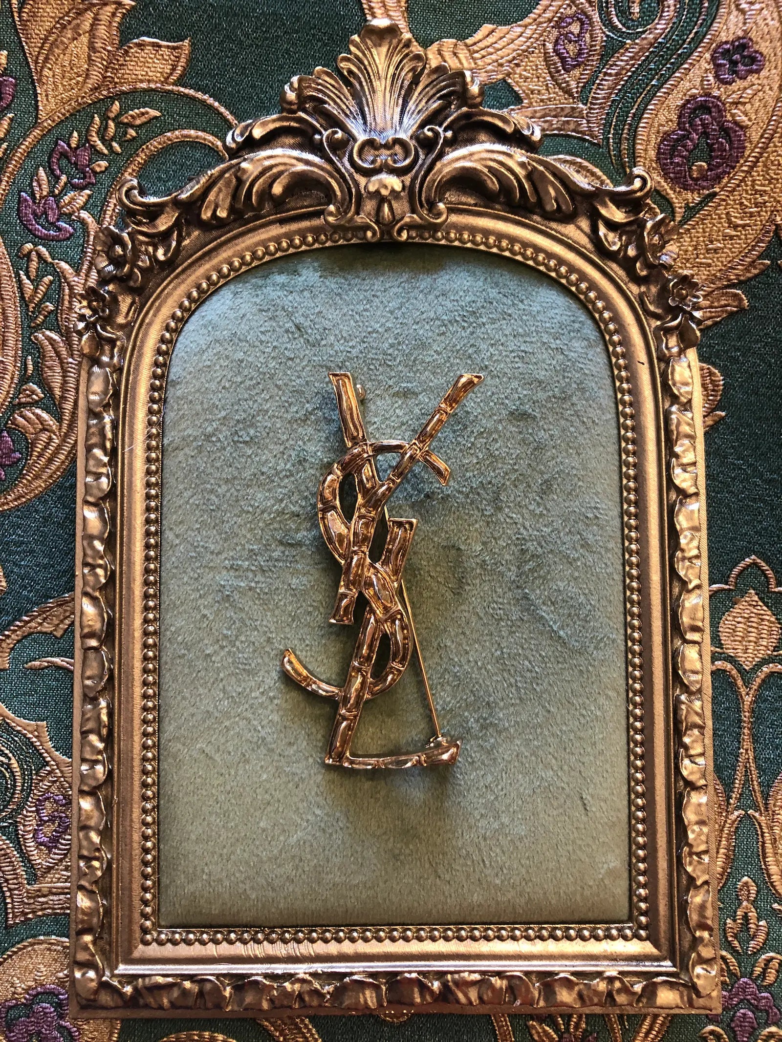 Broochella Yves Saint Laurent costume gold tone metal logo brooch. It is displayed on a green velvet tray with a gold frame. The background is green and gold damask wallpaper