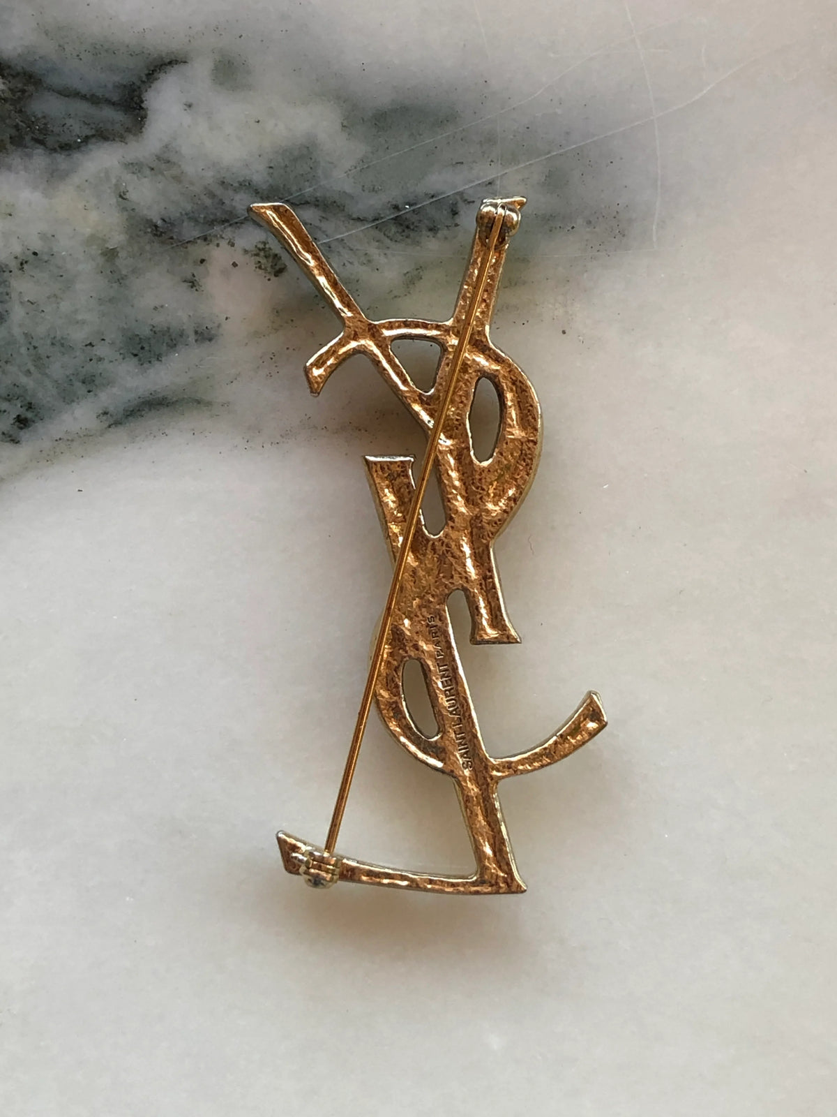 The back view of Broochella Yves Saint Laurent costume, gold tone logo brooch. the background is white marble