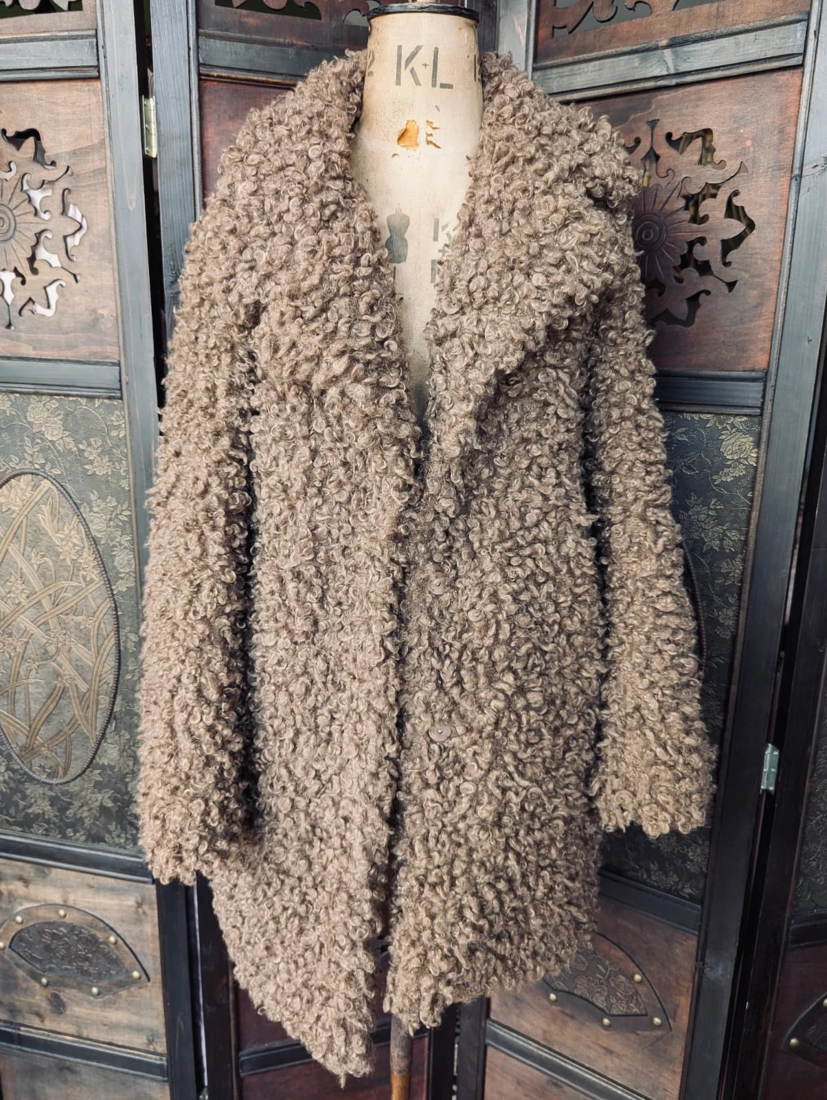 Broochella Zara Brown Teddy Faux fur coat. It is pictured on a vintage mannequin The background is an ornate, vintage, dark wood room divider.