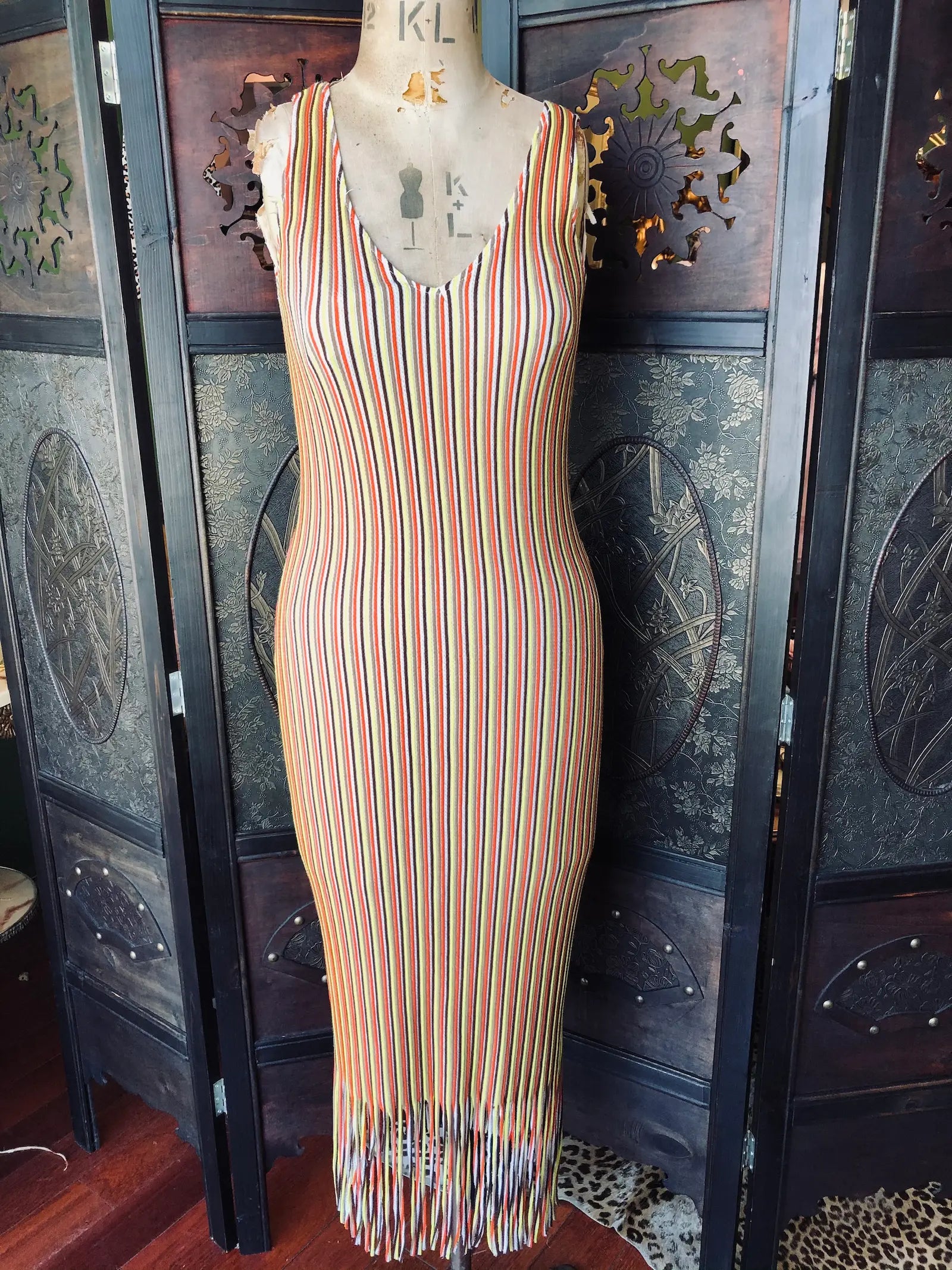 Broochella Zara size small, brown, orange, yellow and white stripe midi dress, with  V-neckline, sleeveless, slim silhouette and fringe hem detail. It is pictured on a vintage mannequin. The background is an ornate, vintage, dark wood room divider. 