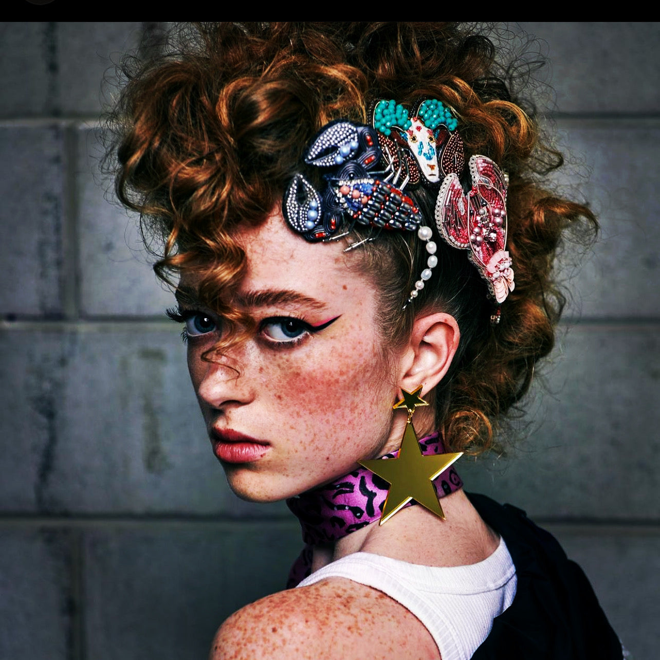 Red, curly headed model with Broochella zodiac beaded brooches in her hair. She is looking over her shoulder to face the camera. The background is a grey brick wall.