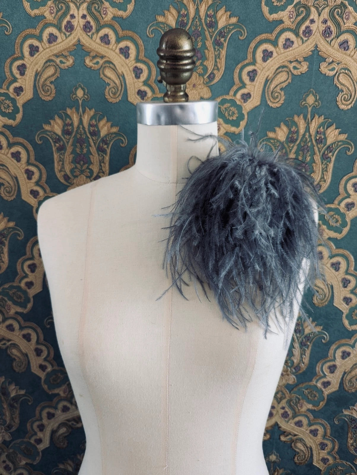 Broochella le pouffe handmade designer feather brooch in silver.  It is pinned to a mannequin. The background is green and gold damask wallpaper