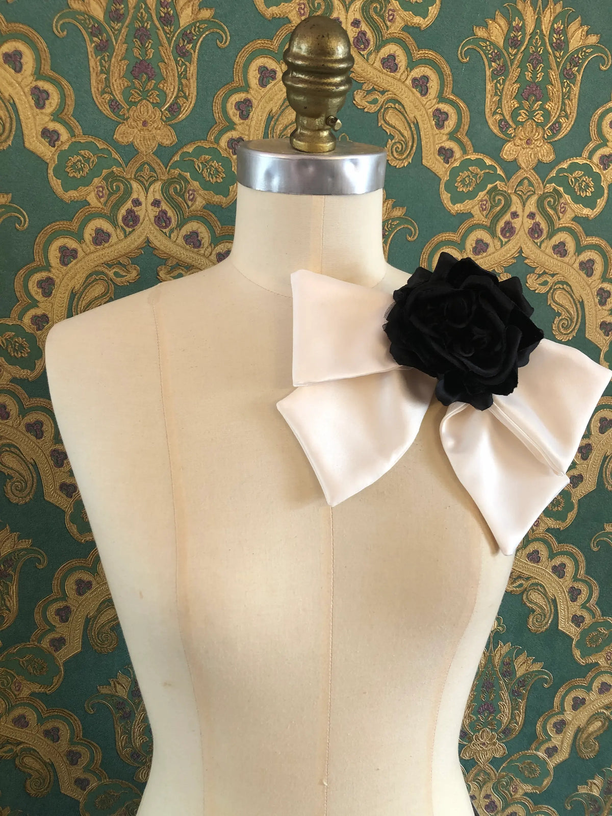 Broochella Dolce Amore Bow Brooch in ivory silk with a dramatic black floral centre, styled on a mannequin against ornate green and gold vintage wallpaper.