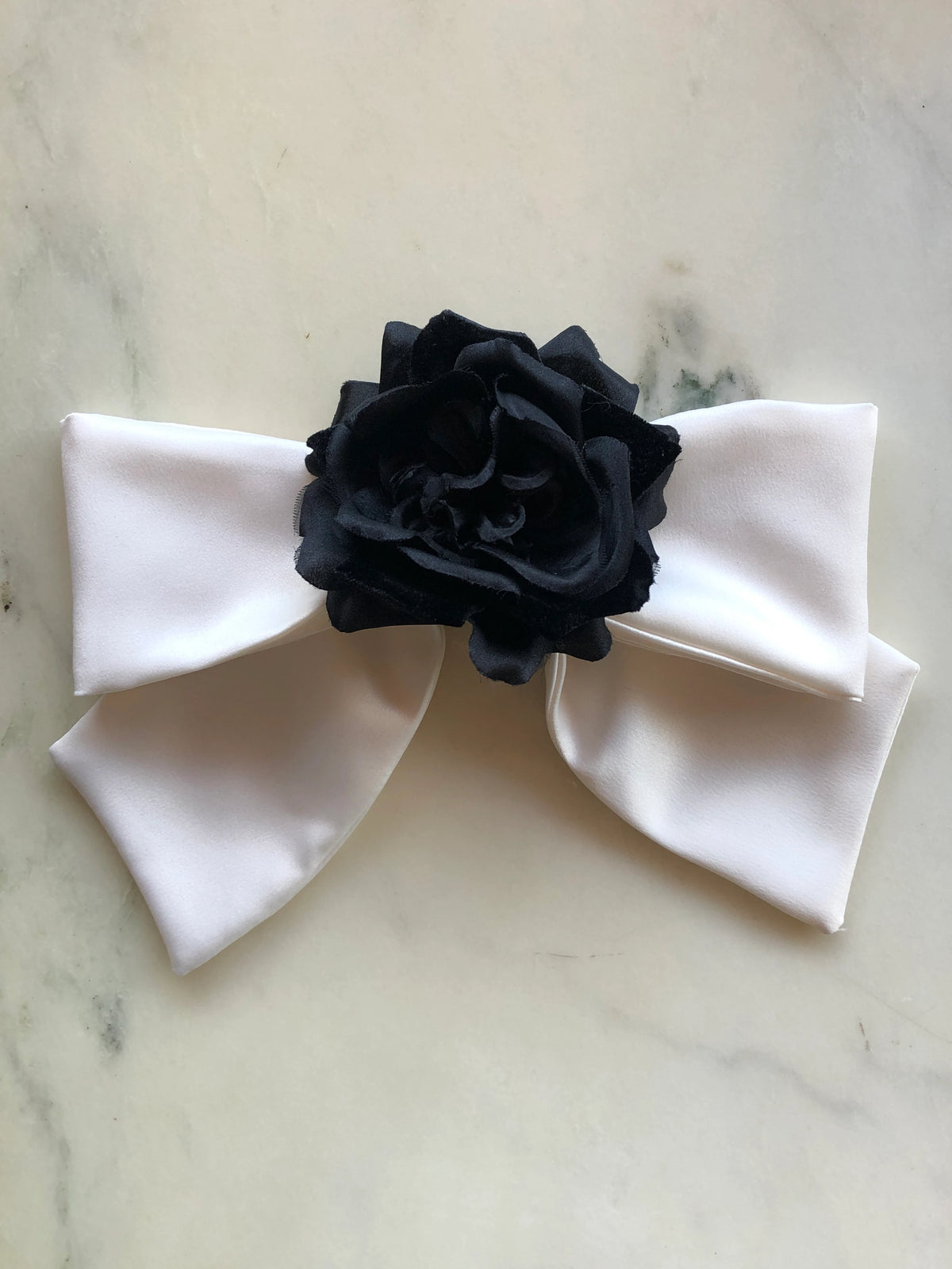 Broochella Dolce Amore Bow Brooch in ivory silk with a statement black floral centre, photographed on a white marble background.