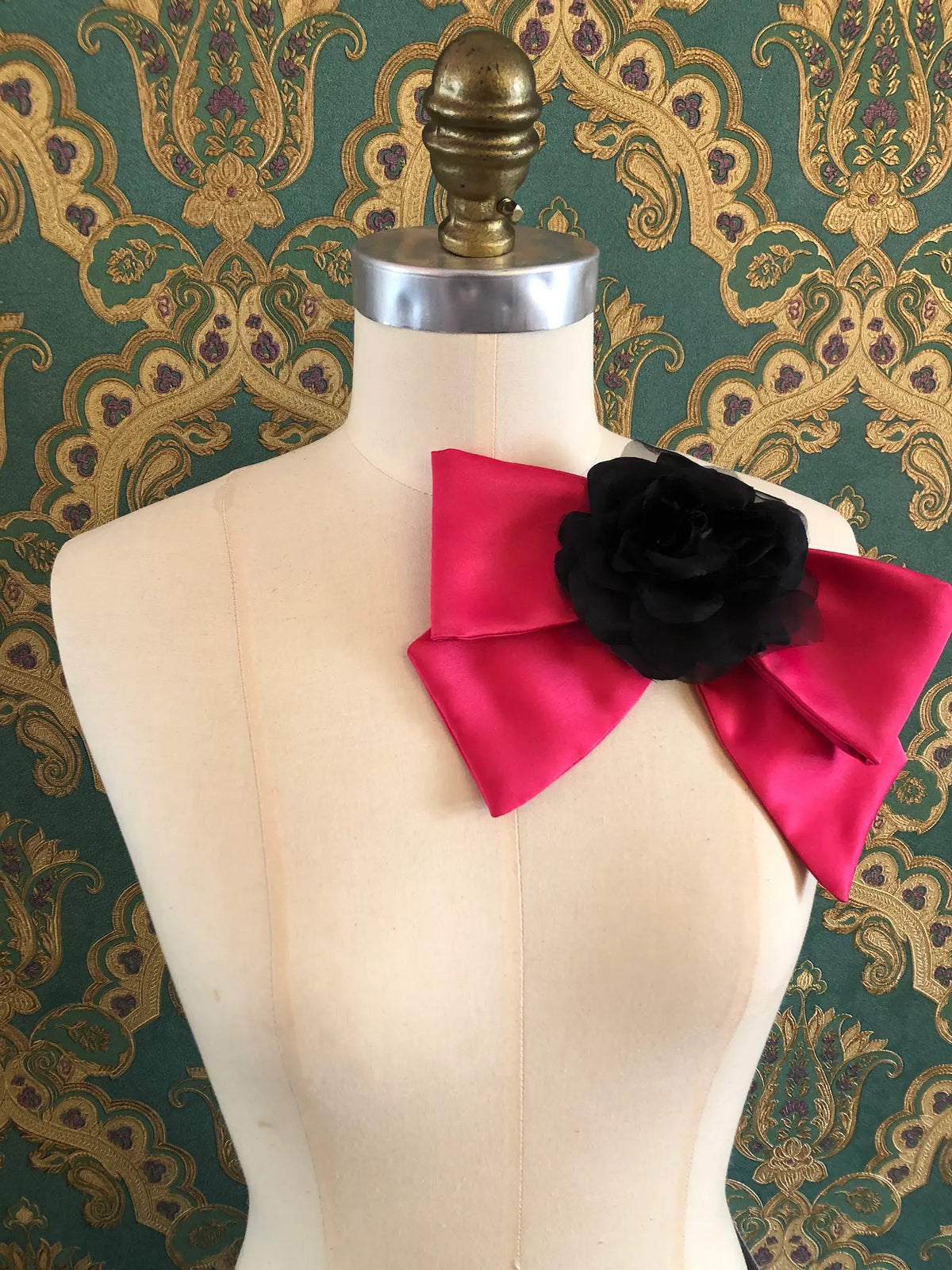 Broochella Dolce Amore Bow Brooch in vibrant pink silk with a dramatic black floral centre, styled on a mannequin against ornate green and gold vintage wallpaper.