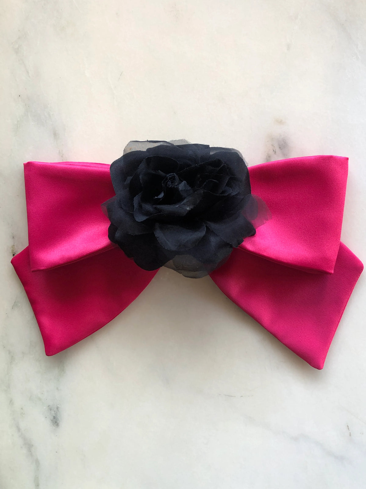 Broochella Dolce Amore Bow Brooch in vibrant pink satin with a statement black floral centre, photographed on a white marble background.