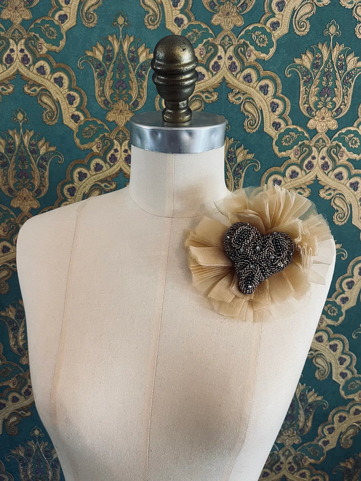 Fiore del Cuore flower brooch in brown, featuring an intricately hand-beaded bronze and black heart surrounded by soft dark brown tulle, displayed on a cream dress form against ornate teal, gold and purple patterned wallpaper.