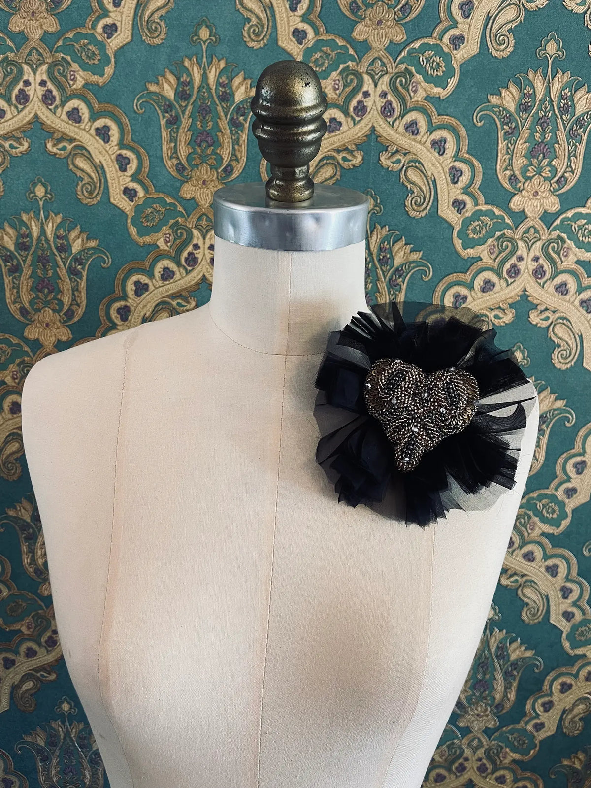 Fiore del Cuore flower brooch in black, featuring an intricately hand-beaded bronze and black heart surrounded by layers of black tulle, displayed on a cream dress form against ornate teal, gold and purple patterned wallpaper.