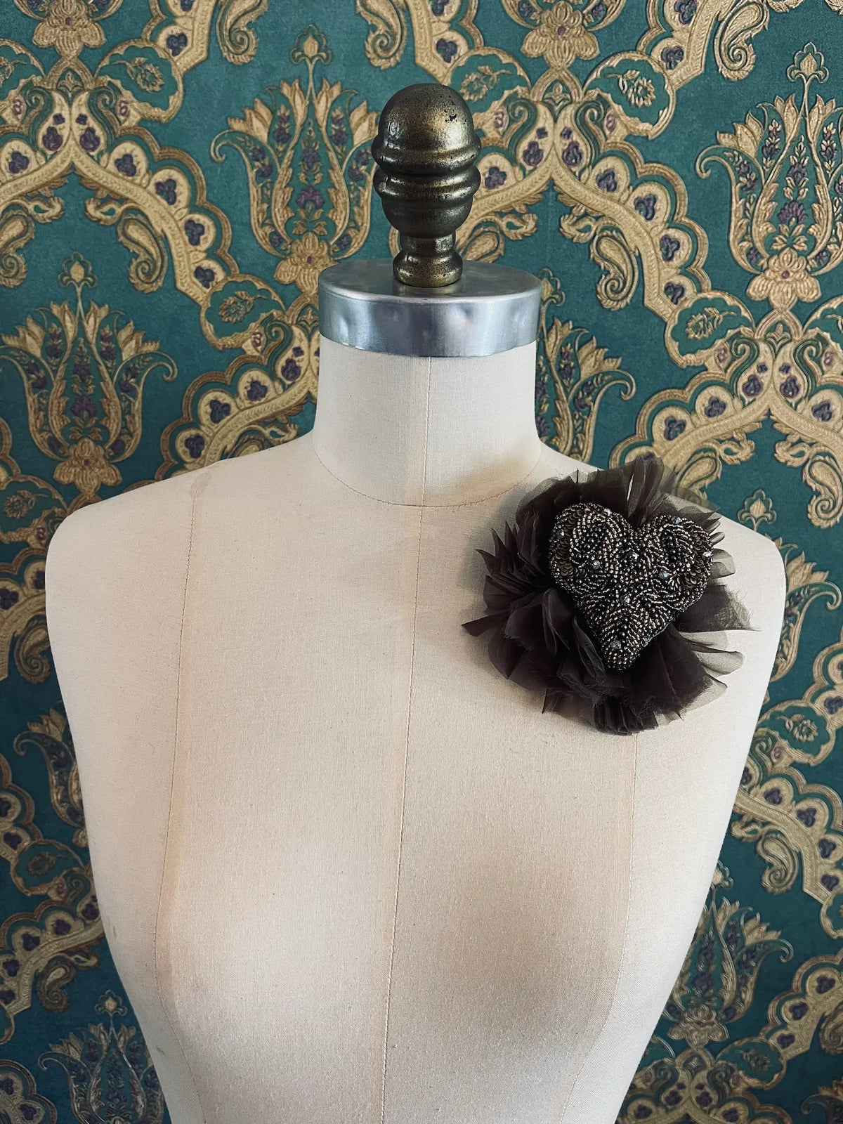 Fiore del Cuore flower brooch in brown, featuring an intricately hand-beaded bronze and black heart surrounded by soft dark brown tulle, displayed on a cream dress form against ornate teal, gold and purple patterned wallpaper.