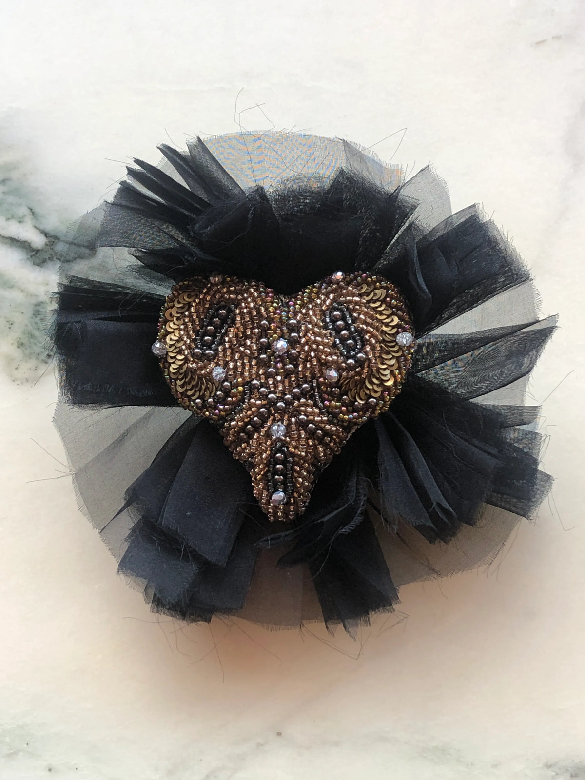 Fiore del Cuore flower brooch featuring a bronze, gold and black hand-beaded heart with sequins and crystals, set against layers of black tulle and fabric petals, photographed on a white marble background with soft grey veining.