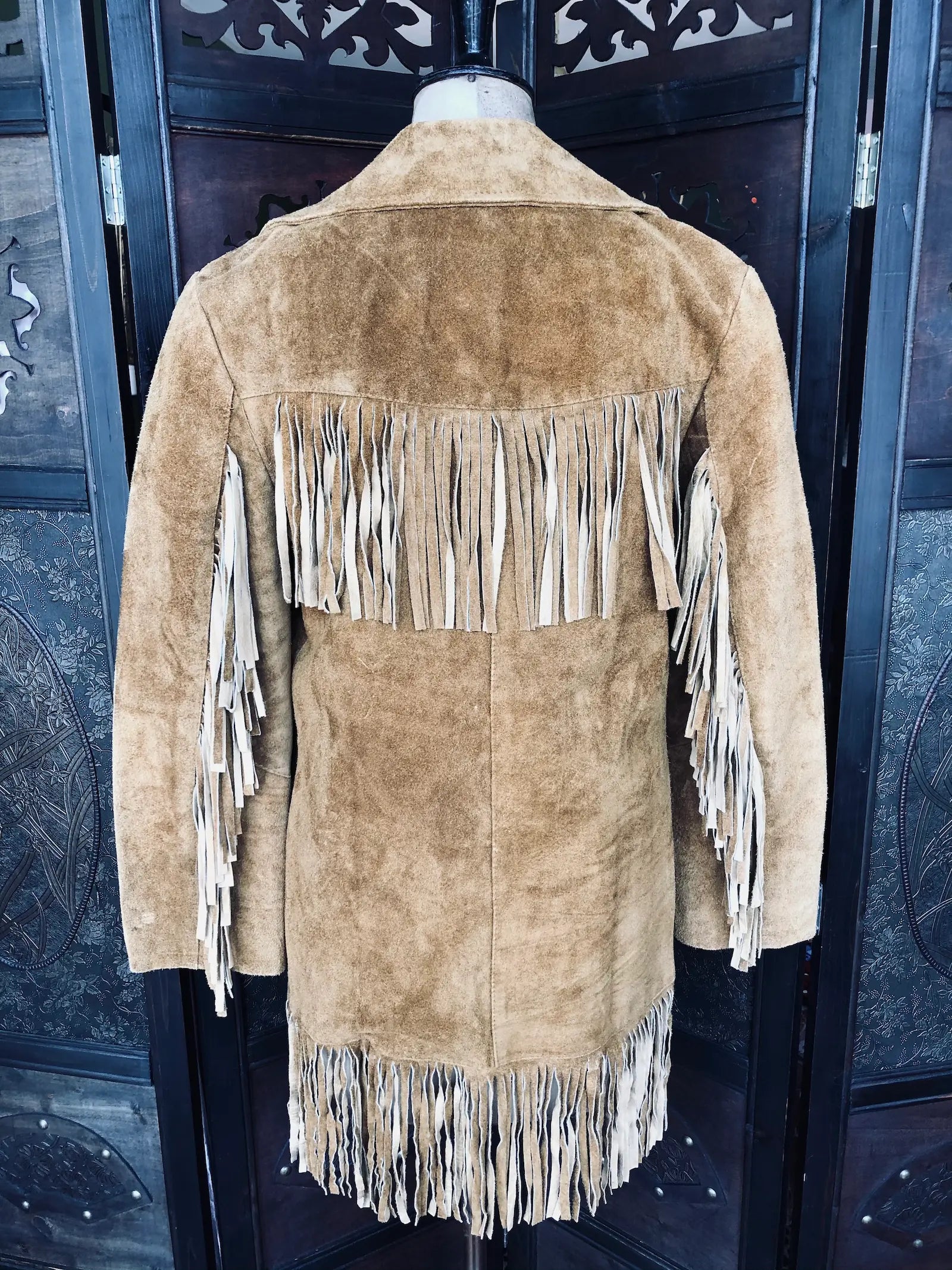 Broochella, The Stylist's Closet Jhill Hats & Clothing Suede Fringe Jacket – Size Small in light brown-pictured from the back.. It is pictured on a mannequin. The background is an ornate, vintage, brown wooden room divider. 
