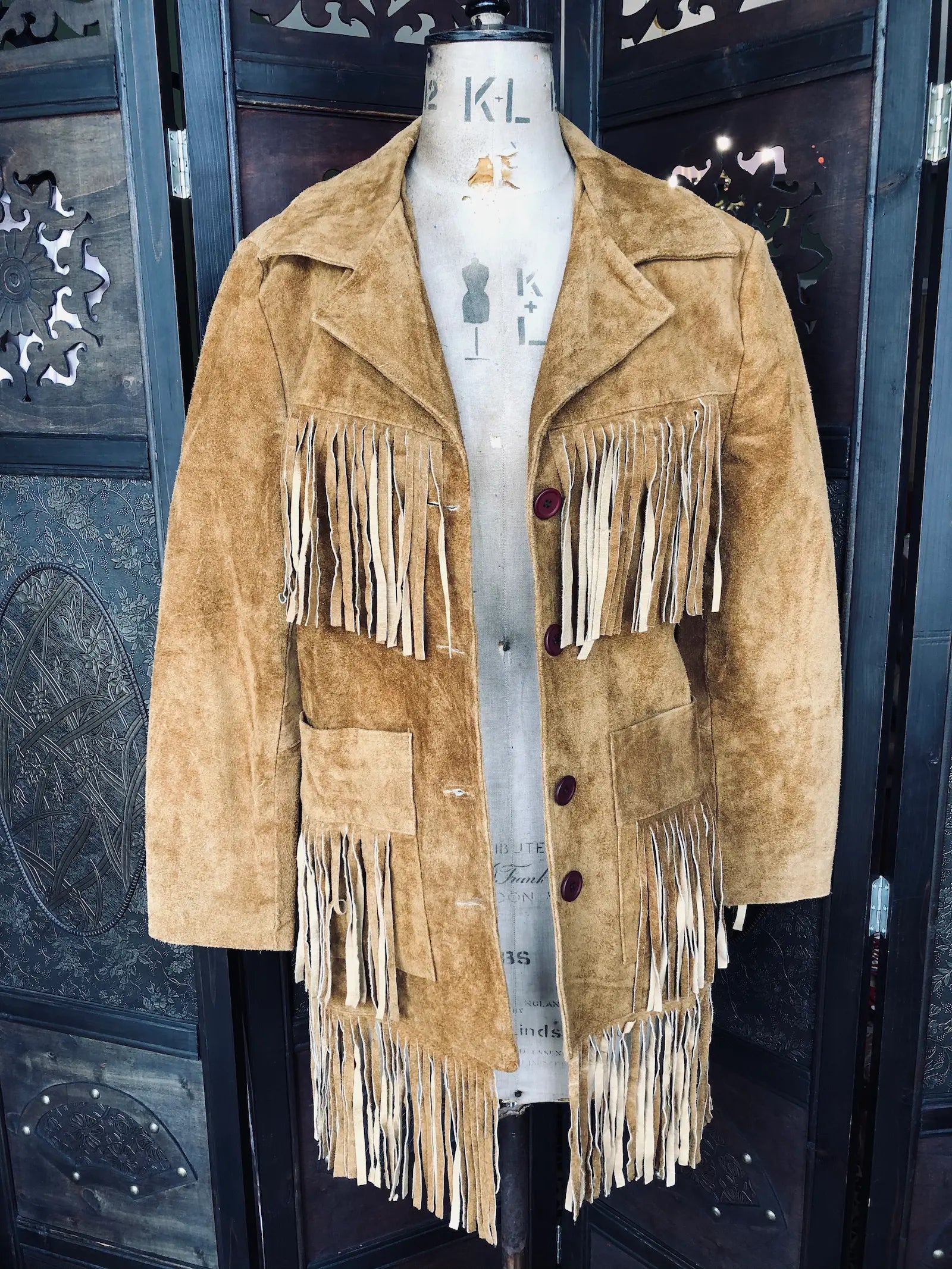Broochella, The Stylist's Closet Jhill Hats & Clothing Suede Fringe Jacket – Size Small in light brown. It is pictured on a mannequin. The background is an ornate, vintage, brown wooden room divider. 