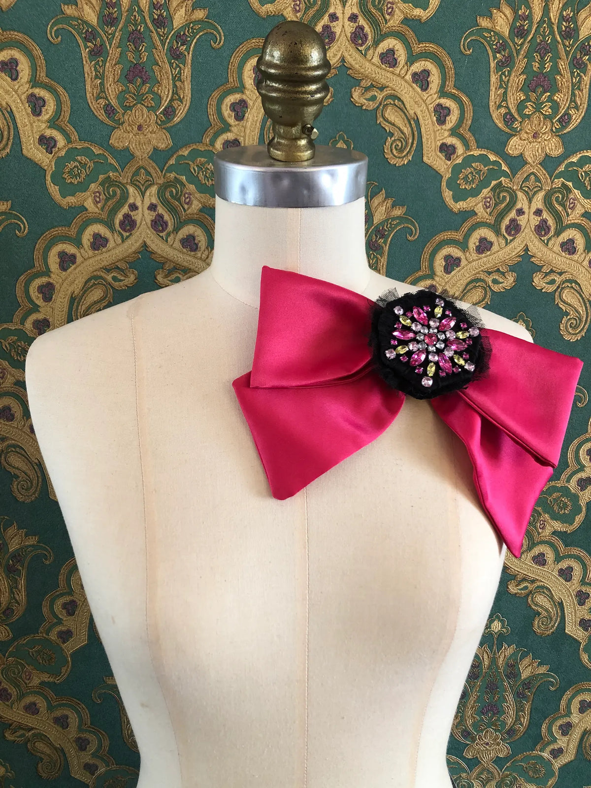 Broochella La Dolce Diva Bejewelled Bow Brooch in vibrant fuchsia satin with a black centre embellished with pink, green and clear crystals, styled on a mannequin.