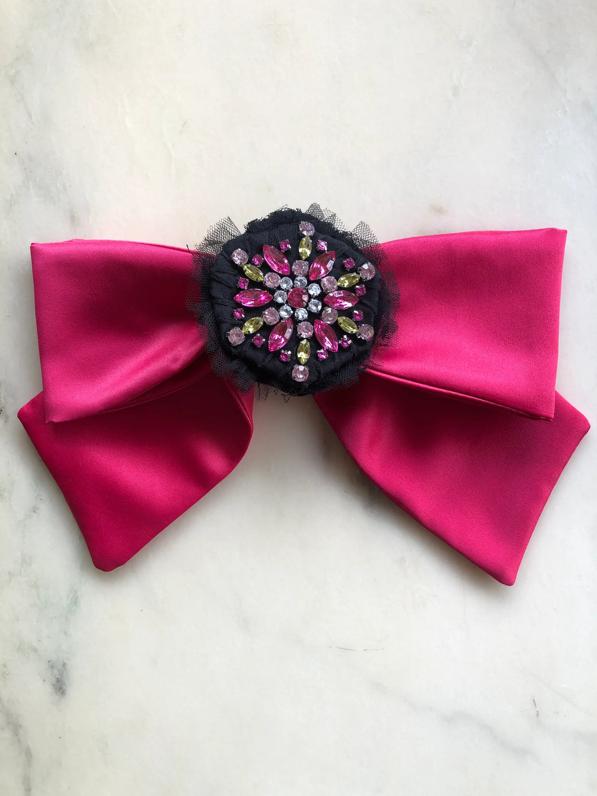 Broochella La Dolce Diva Bejewelled Bow Brooch in vibrant fuchsia satin with an ornate black centre embellished with pink, green and clear crystals, photographed on a white marble background.