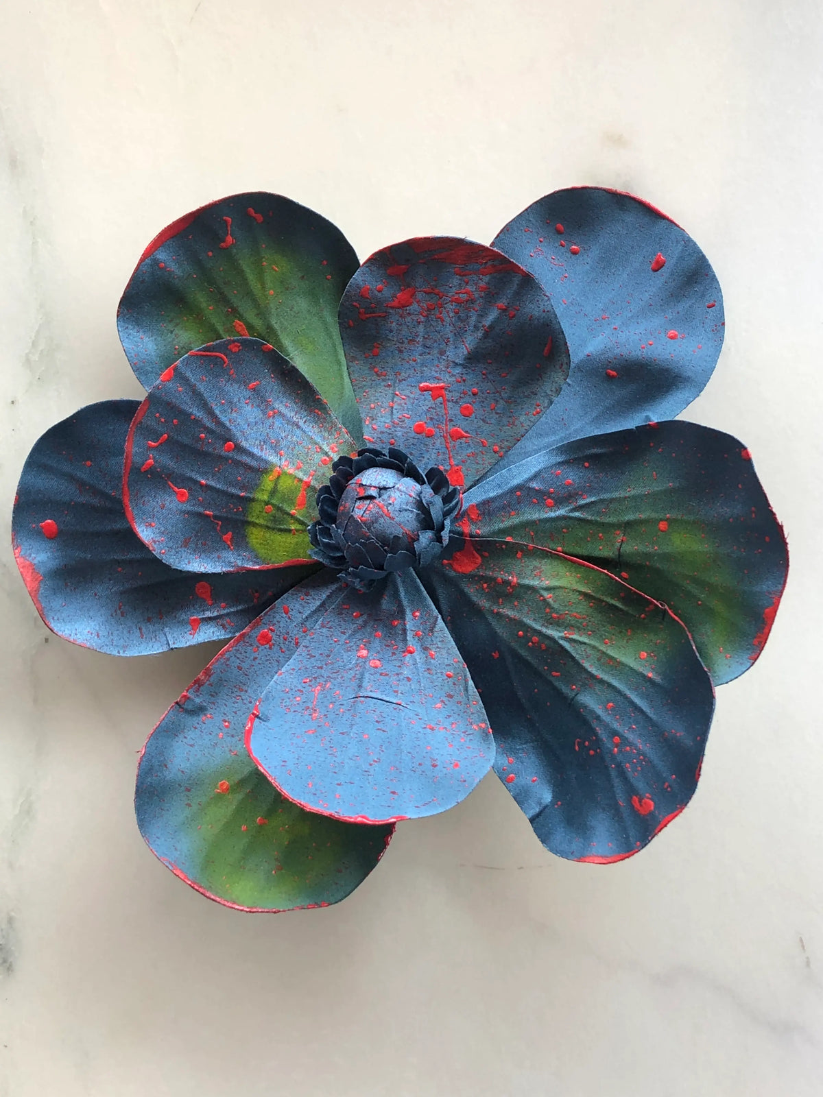 Handmade Broochella Magnolia d’Arte designer flower brooch with oversized sculptural blue silk petals, green painterly accents and vivid red paint splatter detailing, photographed on white marble.