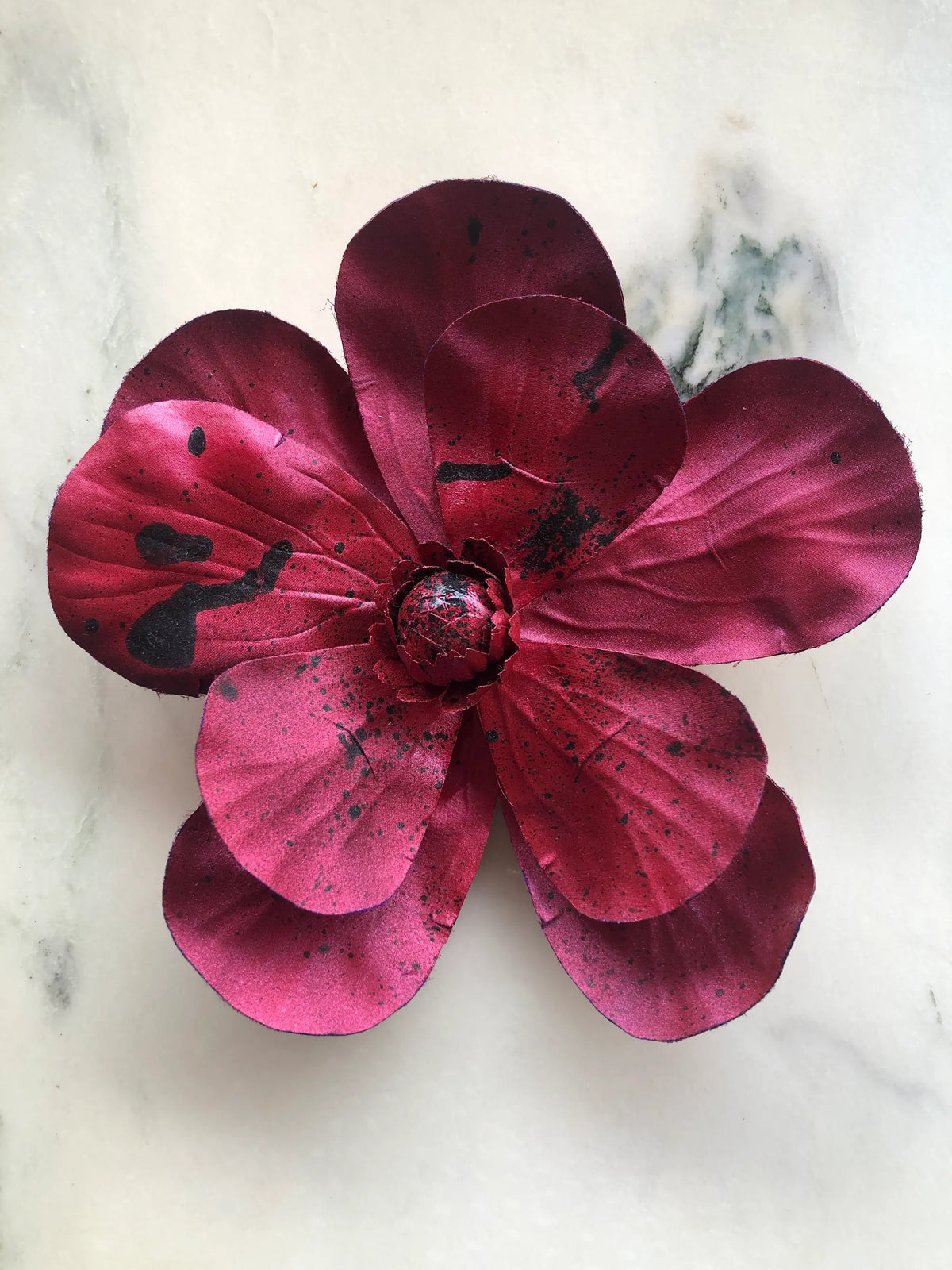 Handmade Broochella Magnolia d’Arte designer flower brooch in rich burgundy silk with sculptural oversized petals and artistic black paint splatter detailing, photographed on white marble.
