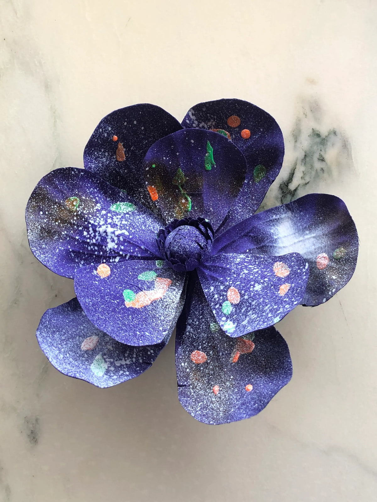 Handmade Broochella Magnolia d’Arte designer flower brooch with oversized sculptural deep purple silk petals, hand-painted white spray detailing and colourful green and orange paint splashes, photographed on white marble.