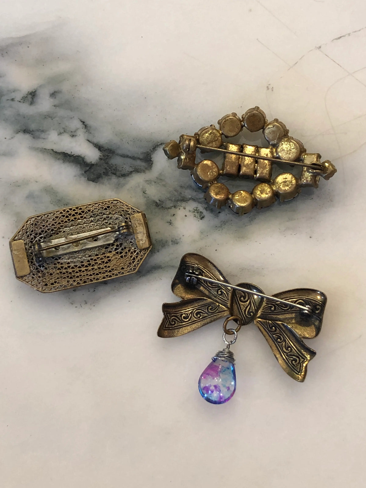 The back view of Broochella Midnight Blue Vintage Brooch Trio featuring blue crystals, a cobalt cabochon and an antique gold bow, displayed on a cream marble background.