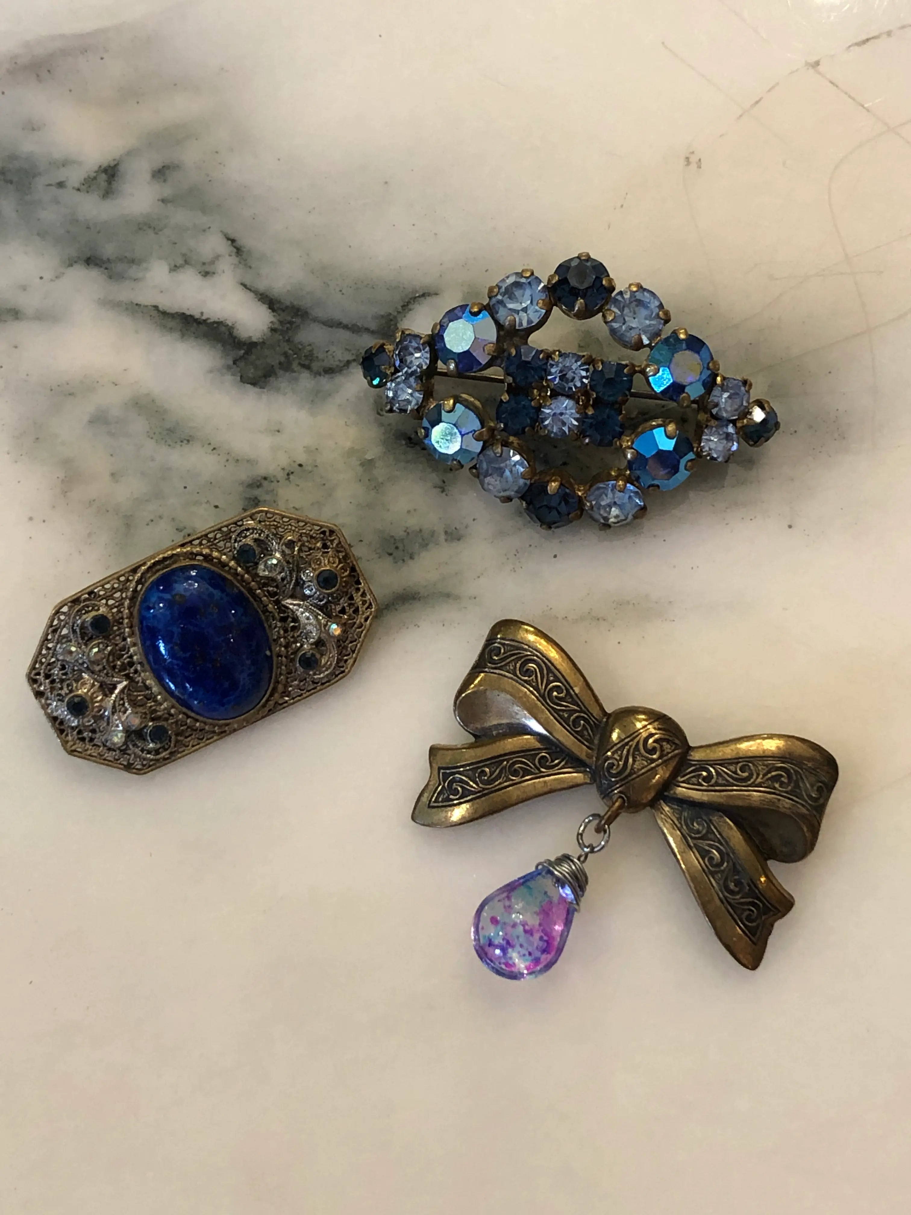 Broochella Midnight Blue Vintage Brooch Trio featuring blue crystals, a cobalt cabochon and an antique gold bow, displayed on a cream marble background.
