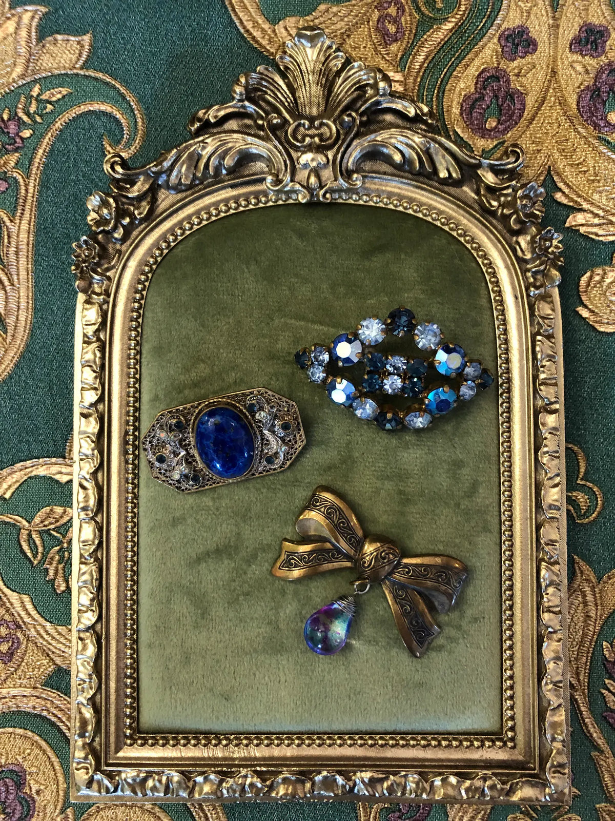 Set of three vintage blue brooches featuring a crystal cluster, cobalt cabochon and antique gold bow with an iridescent teardrop.  It is pictured on a green velvet tray with an ornate gold frame. The background is green and gold damask wallpaper.