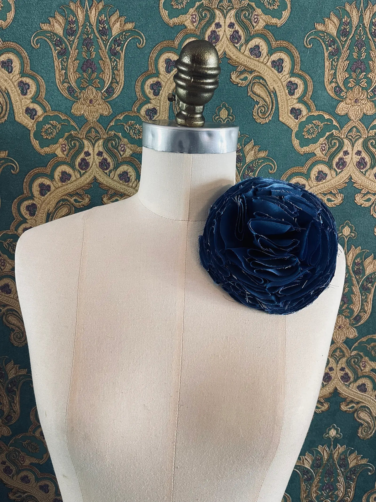 Large Pom Pon Flower Brooch in blue fabric with sculptural layered raw-edged petals, displayed on a cream dress form against ornate teal, gold and purple patterned wallpaper.