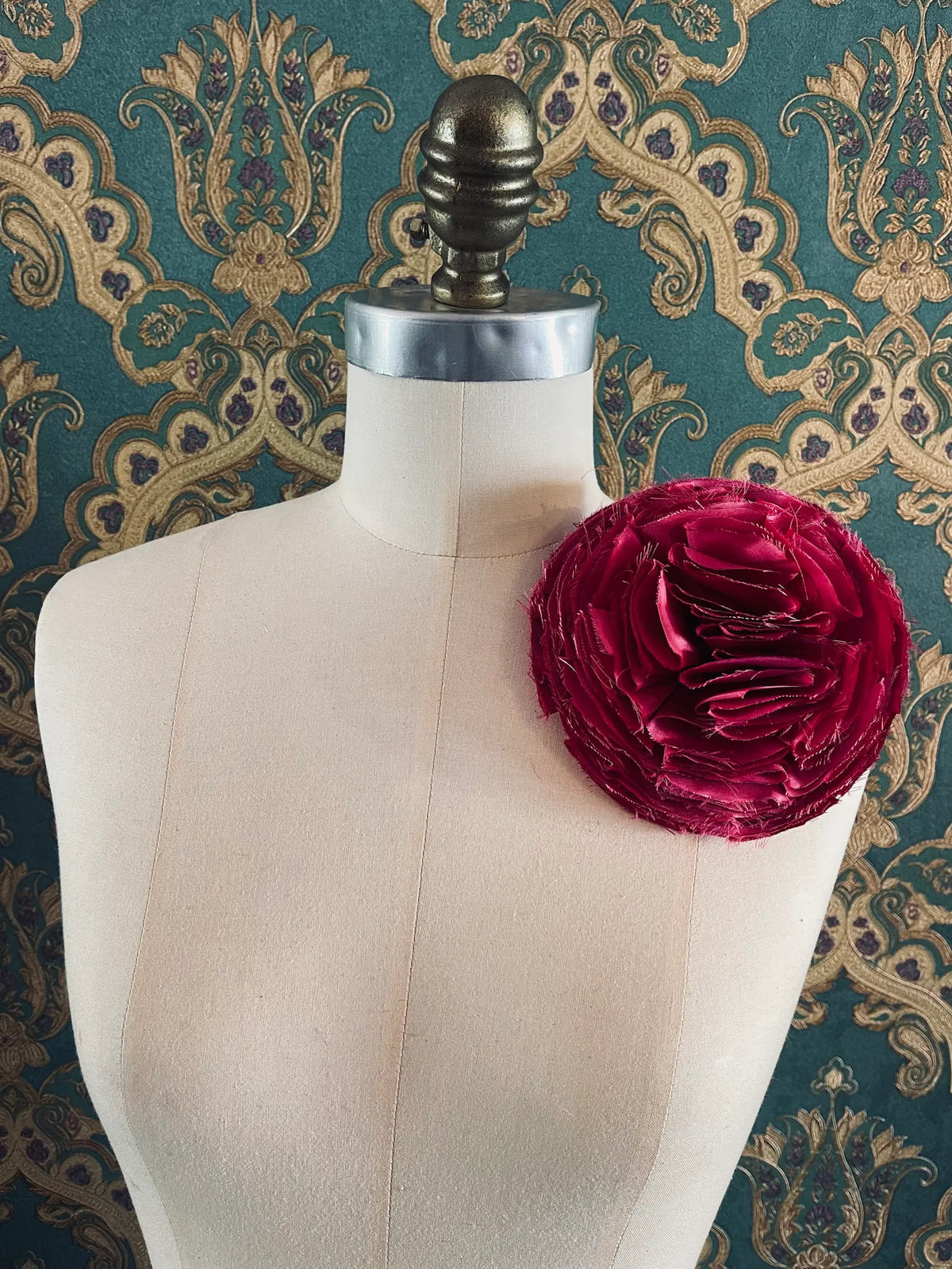 Large Pom Pon Flower Brooch in pink fabric with sculptural layered raw-edged petals, displayed on a cream dress form against ornate teal, gold and purple patterned wallpaper.
