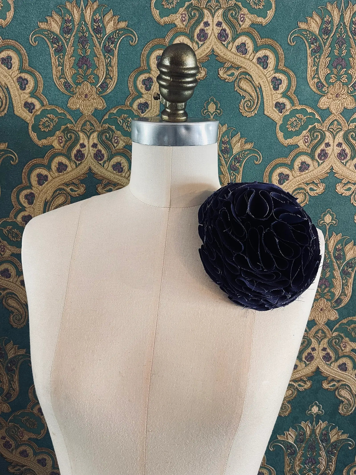 Large Pom Pon Flower Brooch in purple fabric with sculptural layered raw-edged petals, displayed on a cream dress form against ornate teal, gold and purple patterned wallpaper.