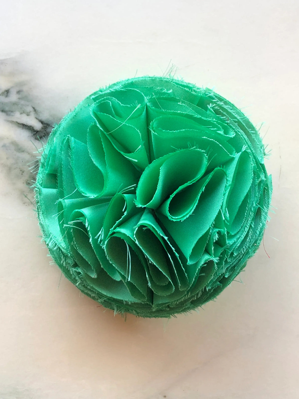 Green Pom Pon small fabric flower brooch with layered raw-edge petals, photographed on a white marble background with soft grey veining.