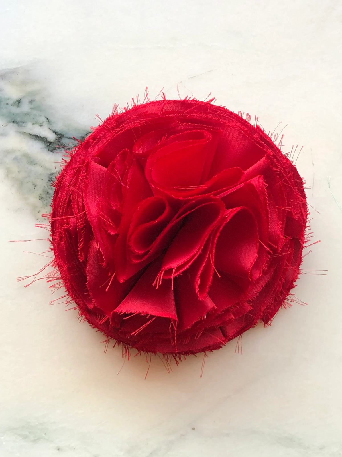 Red Pom Pon small fabric flower brooch with layered raw-edge petals, photographed on a white marble background with soft grey veining.