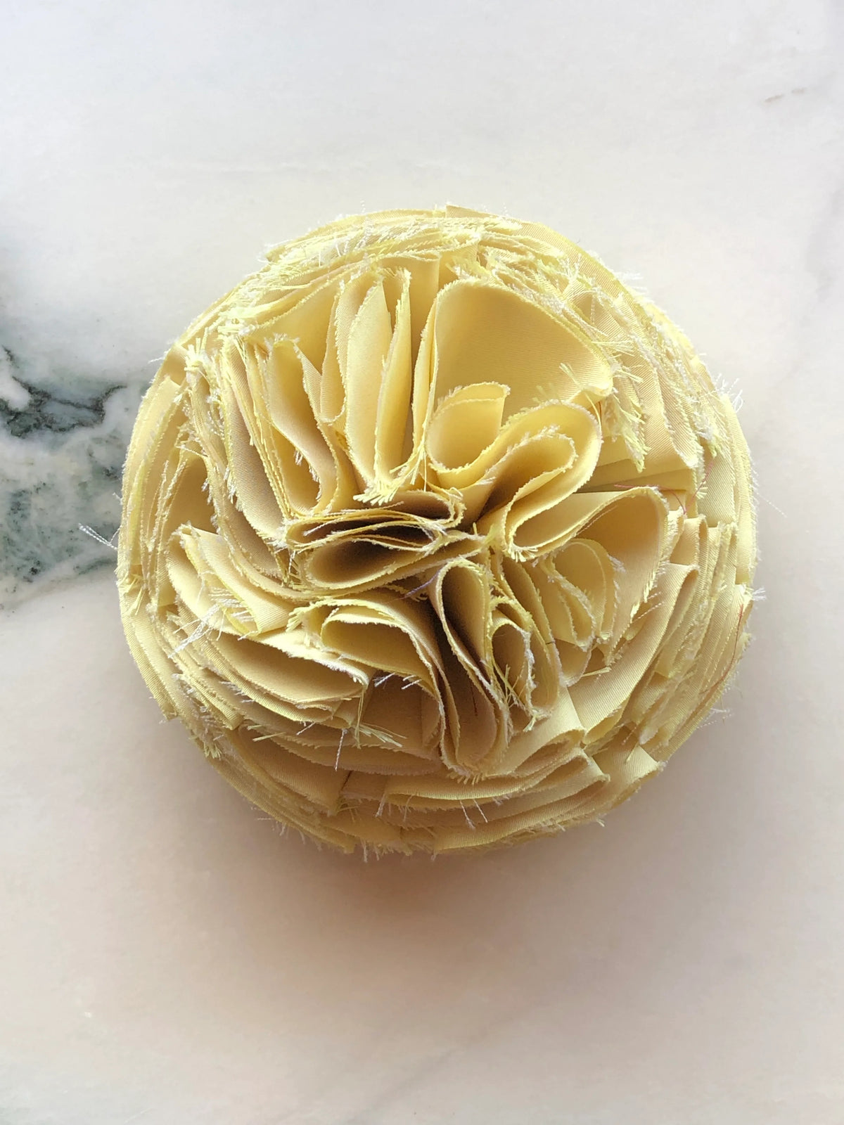 Yellow Pom Pon small fabric flower brooch with layered raw-edge petals, photographed on a white marble background with soft grey veining.