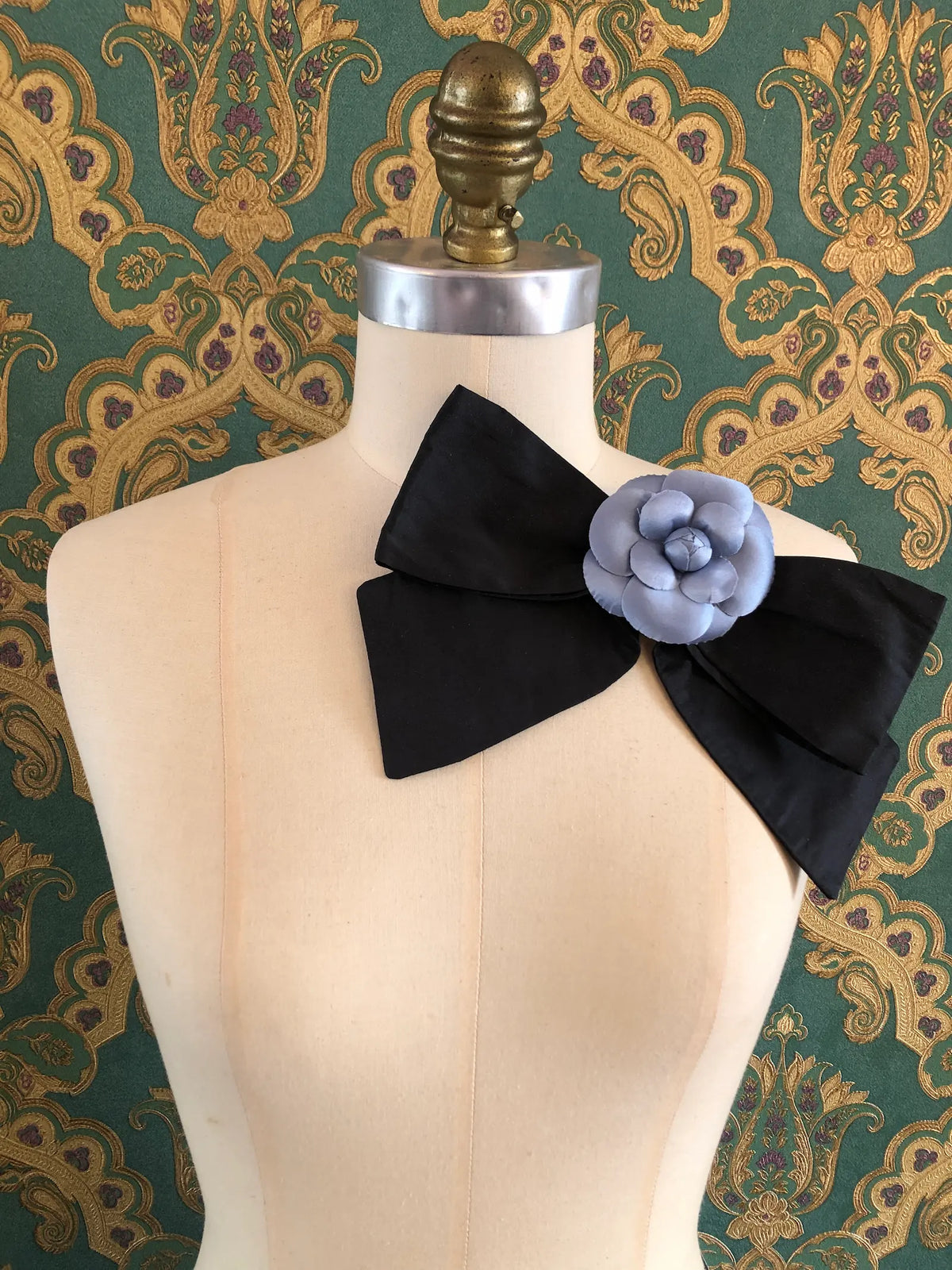 Alt text: Broochella Viva L’Amor Silk Floral Bow Large in black with a powder blue flower centre, styled on a mannequin against ornate green wallpaper.