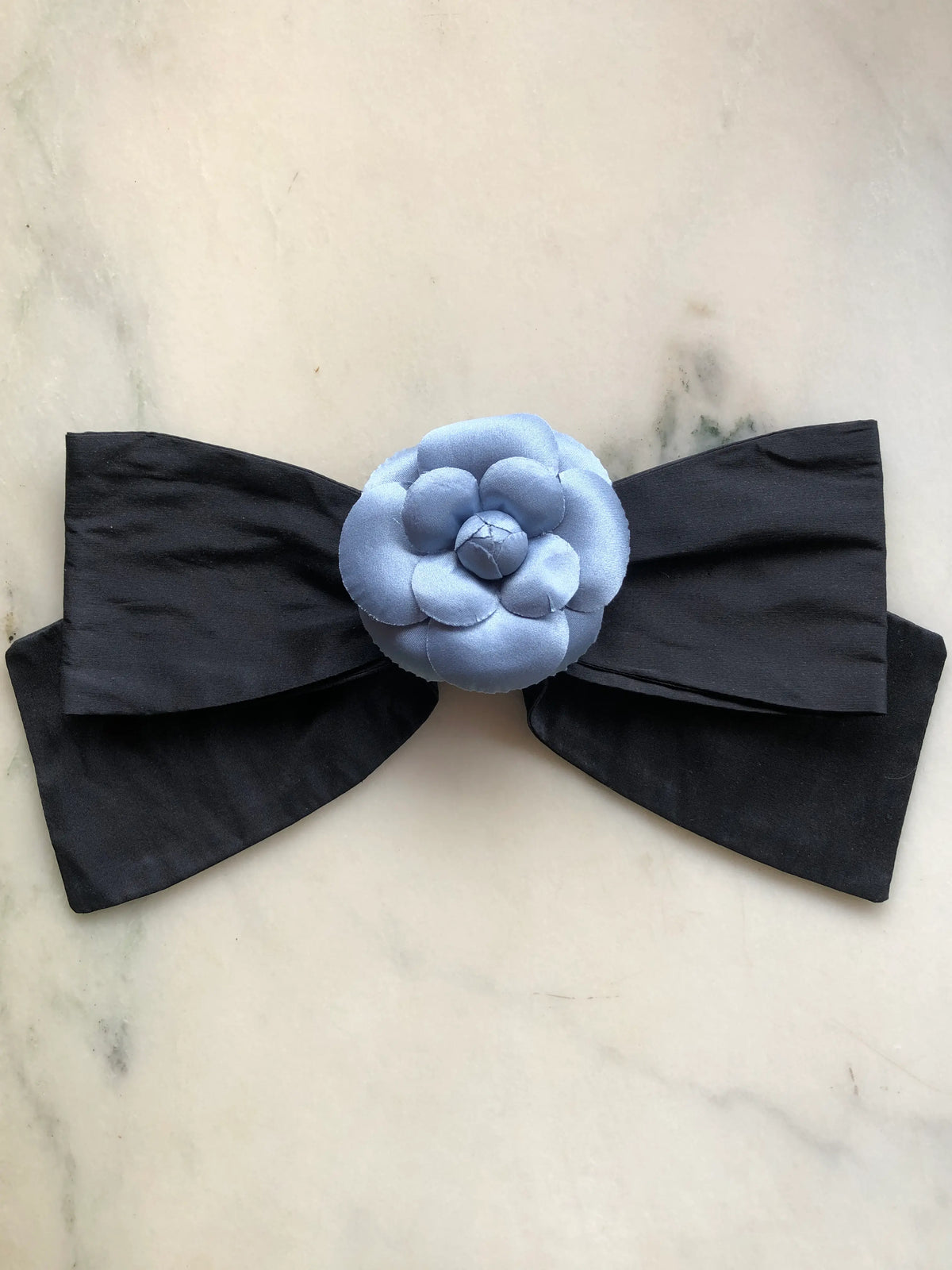Broochella Viva L’Amor Silk Floral Bow Large featuring a black silk bow with a powder blue flower centre, photographed on a white marble background.