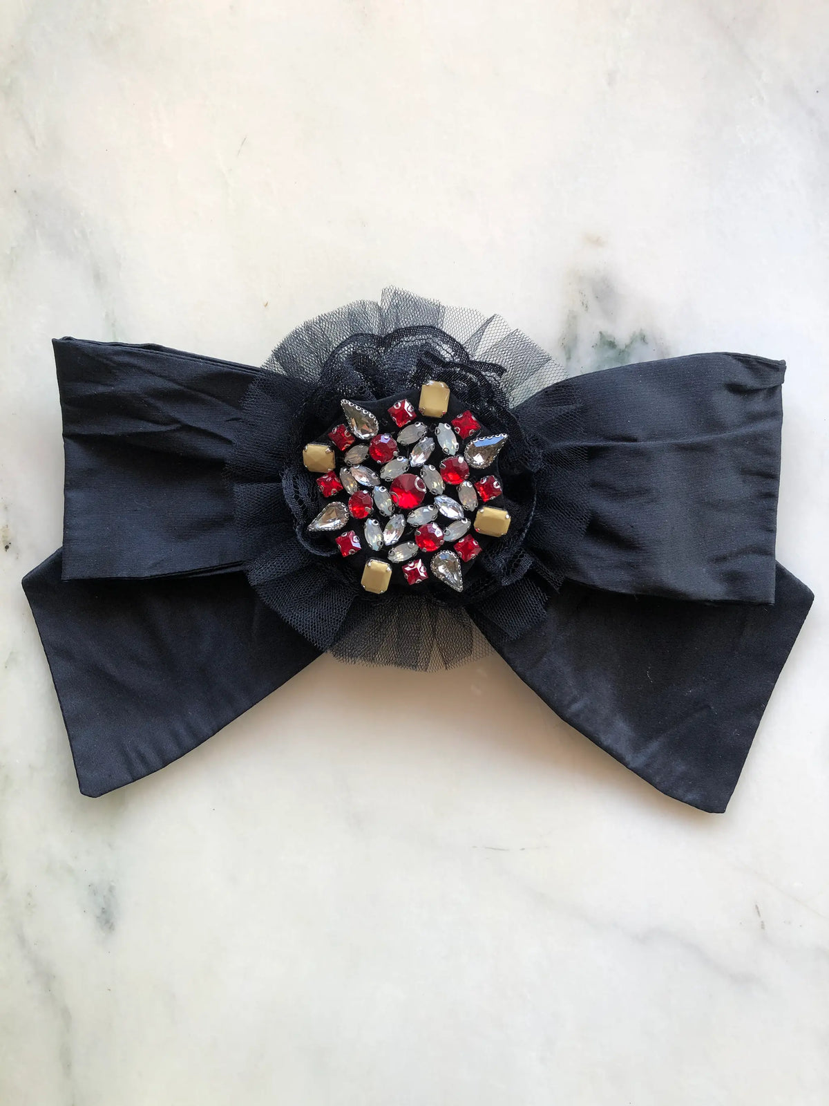 Broochella Viva Loren Bejewelled Bow Brooch in black silk with a tulle-trimmed centre embellished with red, clear and gold crystals, photographed on a white marble background.